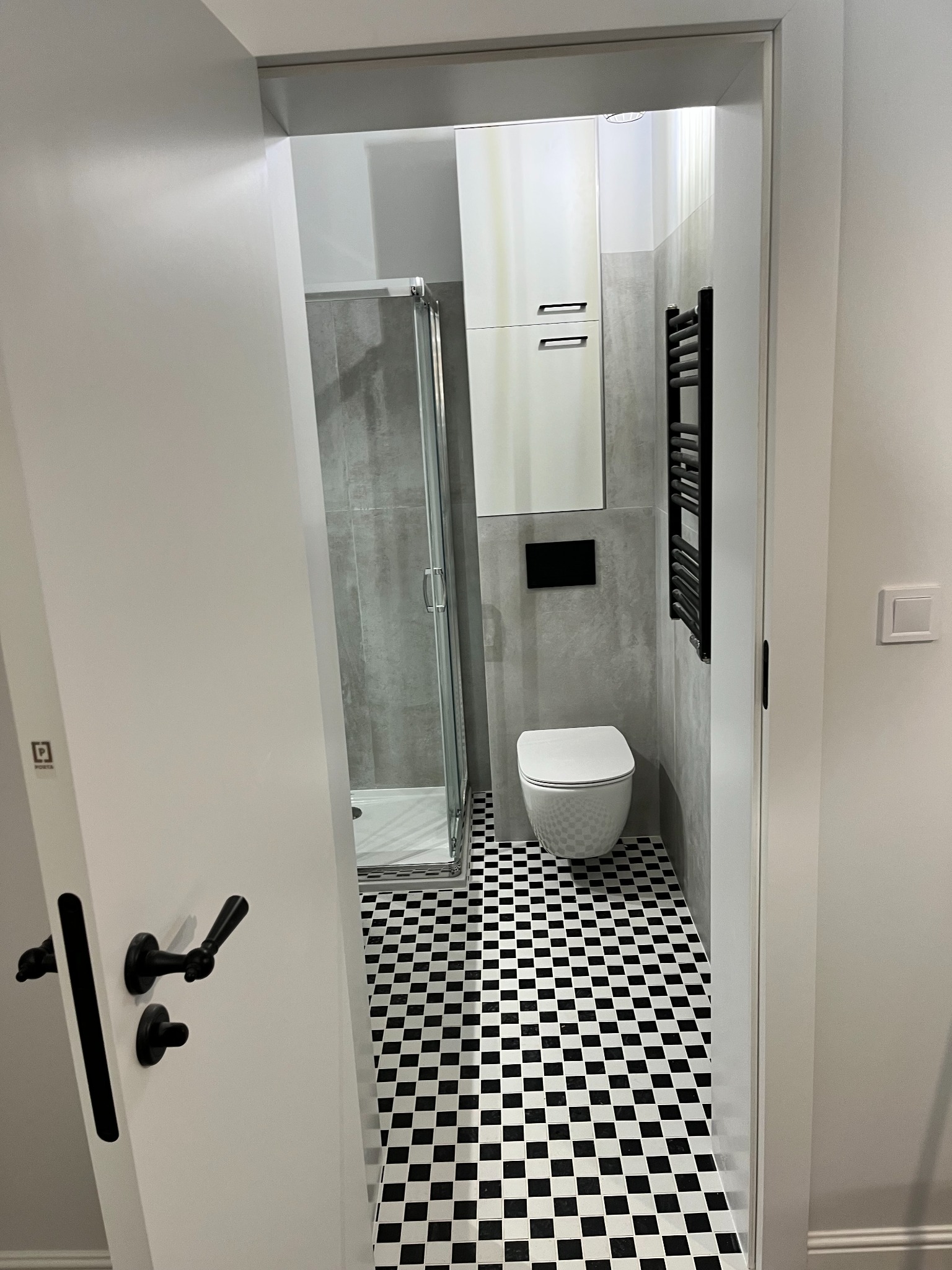 Nowoczesna łazienka z kabiną prysznicową, wiszącą toaletą, grzejnikiem drabinkowym i biało-czarną podłogą w kratkę, widziana zza uchylonych białych drzwi z czarną klamką.