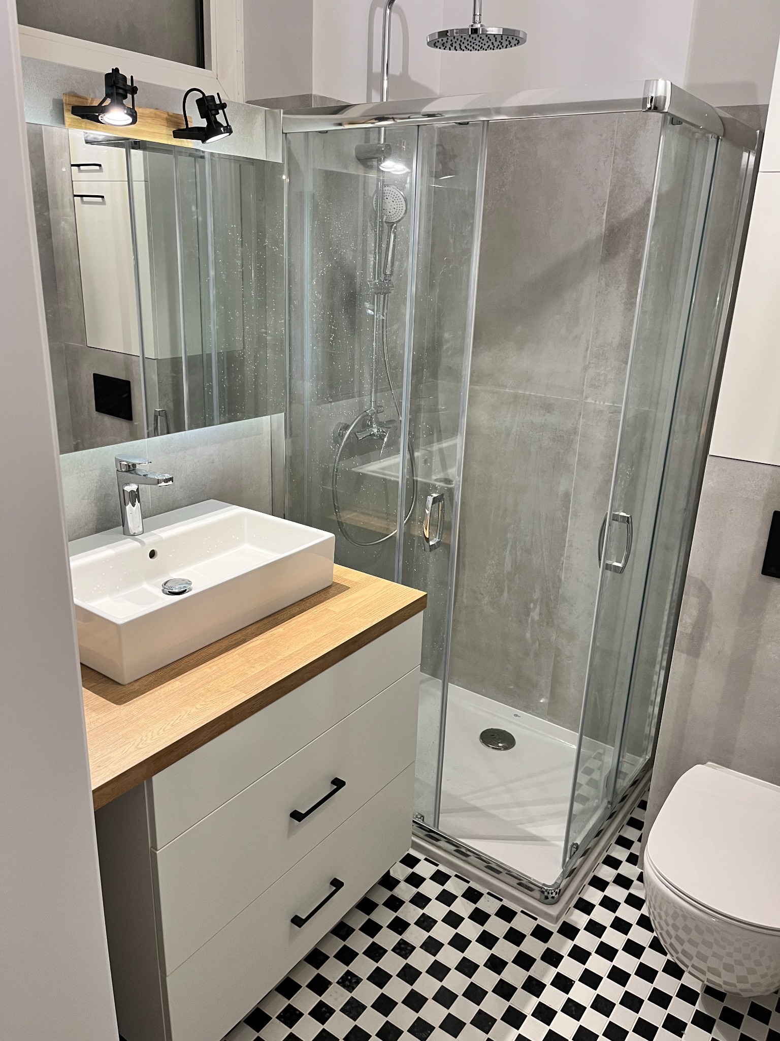 Nowoczesna łazienka z kabiną prysznicową, umywalką nablatową na szafce z drewnianym blatem i czarno-białą podłogą w szachownicę, oświetlenie punktowe nad lustrem.