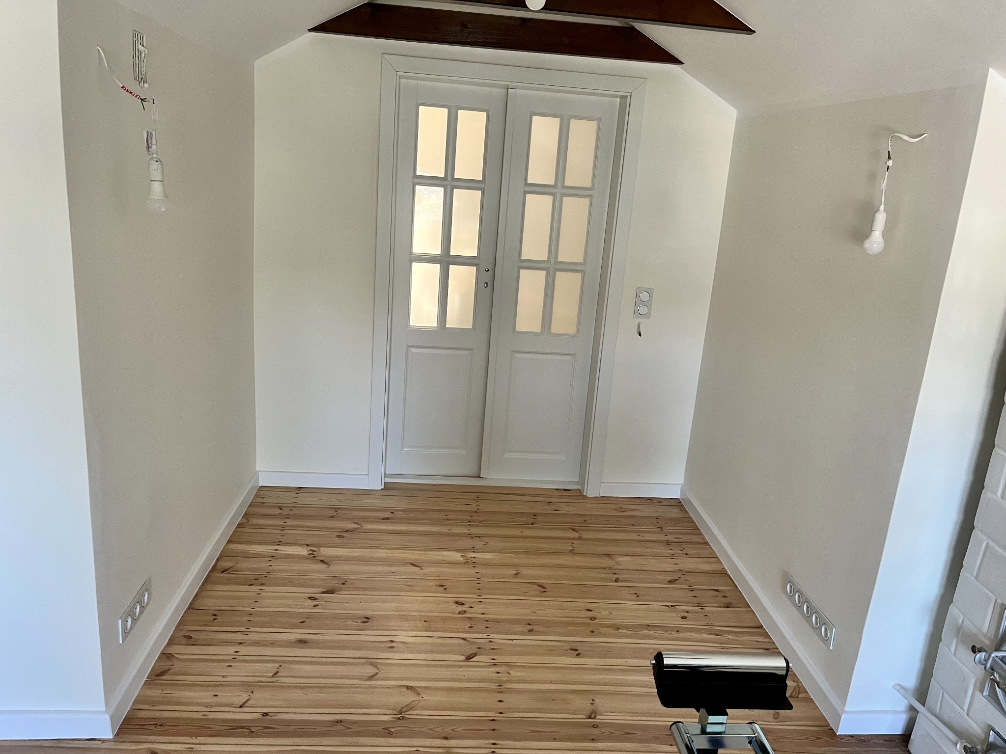 Poddasze po remoncie: świeżo pomalowane białe ściany, odnowiona drewniana podłoga, białe drzwi dwuskrzydłowe z przeszkleniami, widoczne gniazdka elektryczne i prowizoryczne oświetlenie.