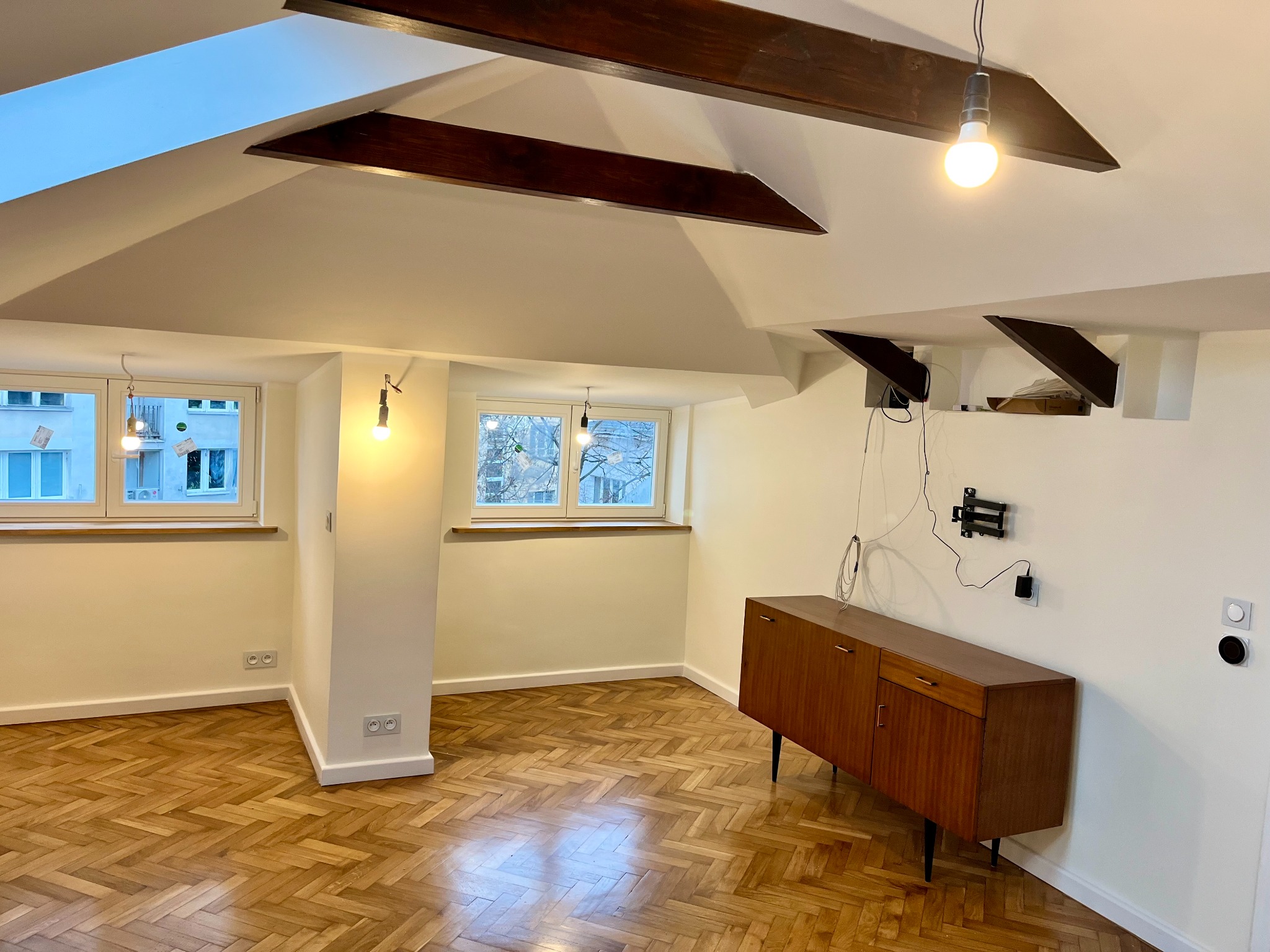 Puste, odnowione wnętrze pokoju z parkietem w jodełkę, drewnianymi belkami stropowymi, wiszącymi lampami na kablach i brązową komodą na tle białych ścian.