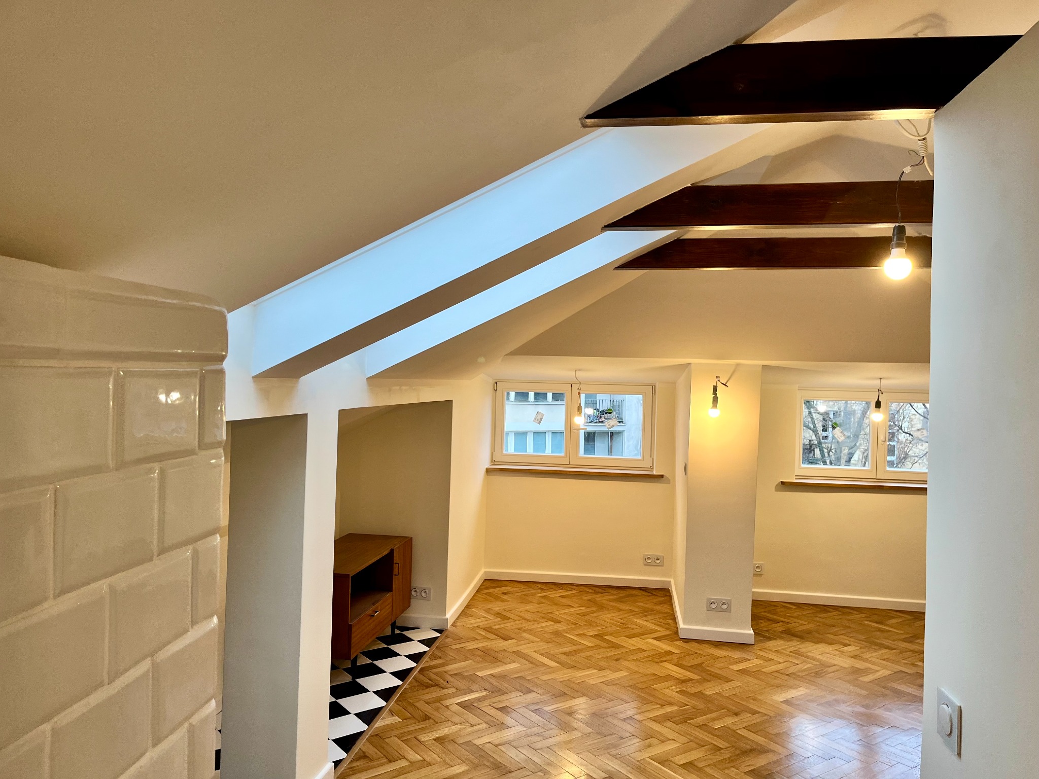 Jasne, przestronne poddasze po remoncie z drewnianą podłogą w jodełkę, białymi ścianami, drewnianymi belkami stropowymi i fragmentem ściany wyłożonej białymi, błyszczącymi kaflami.