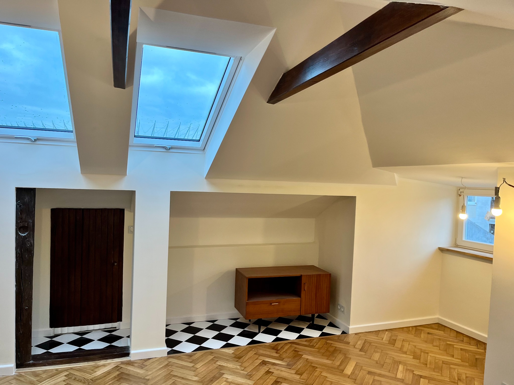 Jasne poddasze z drewnianymi belkami, oknami dachowymi i szachownicową podłogą w niszy z szafką RTV, parkiet w jodełkę.