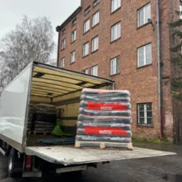 Półciężarówka z otwartą skrzynią załadowana paletą worków z węglem, widoczny wózek paletowy, w tle ceglany budynek z oknami i brama.