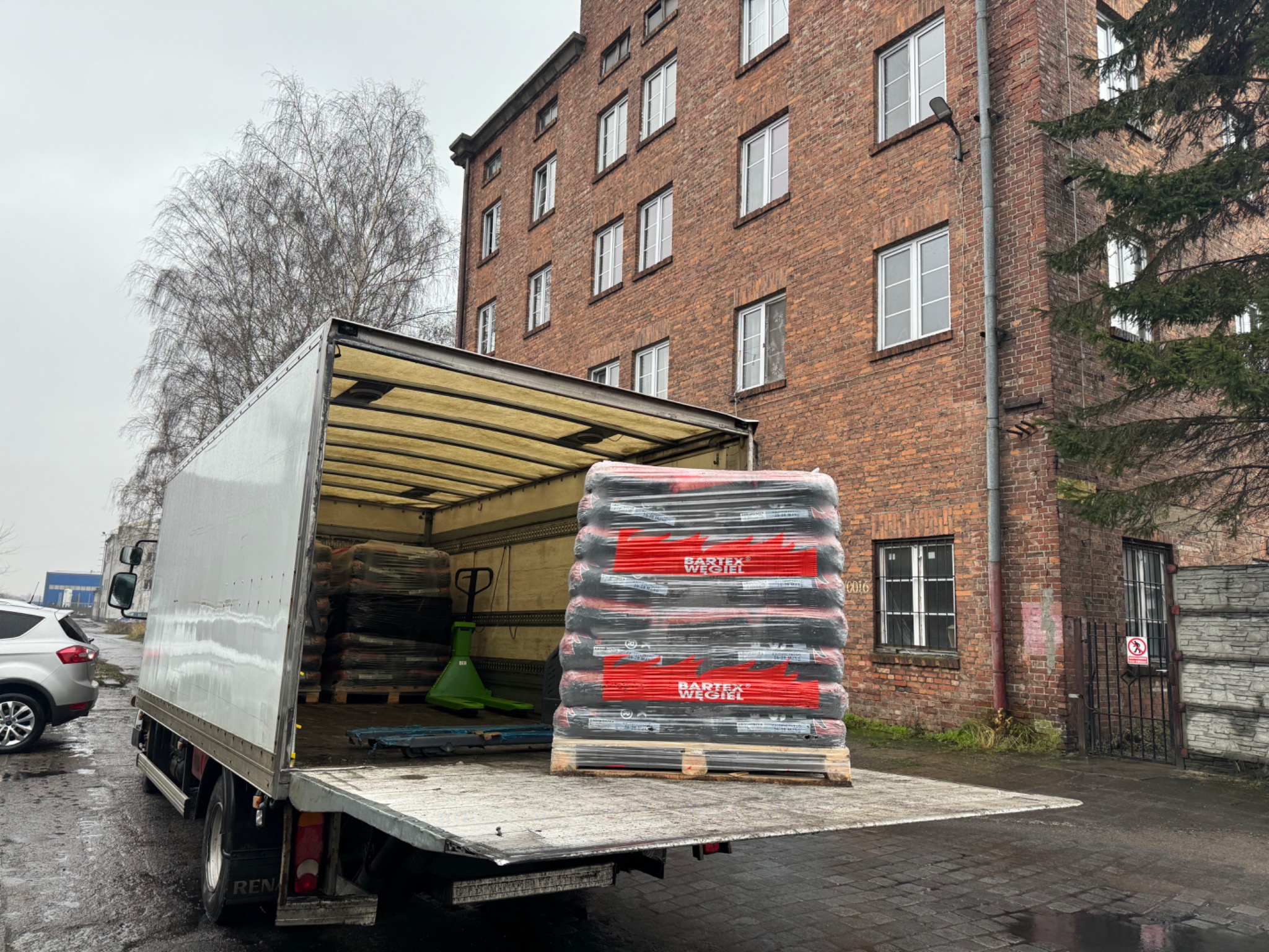 Półciężarówka z otwartą skrzynią załadowana paletą worków z węglem, widoczny wózek paletowy, w tle ceglany budynek z oknami i brama.