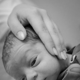 Marlena Jankowska - Czarno-białe zbliżenie niemowlęcia z delikatną dłonią dotykającą jego główki, uwidocznione krótkie włoski dziecka i starannie wypielęgnowane paznokcie dłoni.