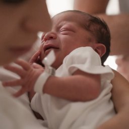 Marlena Jankowska - Noworodek z grymasem na twarzy, trzymany przez osobę w białej koszulce, z widoczną bransoletką identyfikacyjną na rączce dziecka.