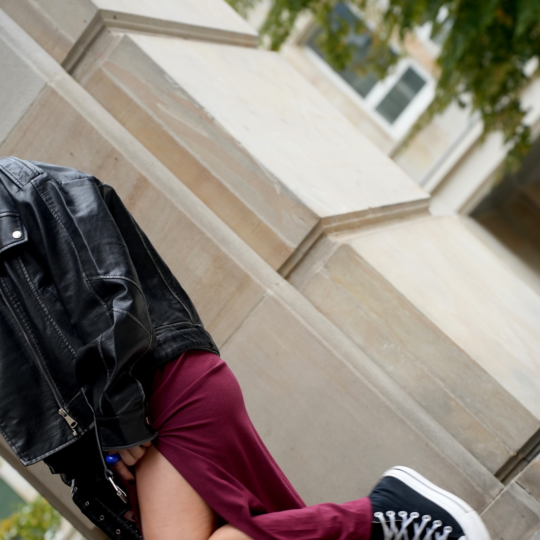 Osoba w czarnej skórzanej kurtce, bordowej sukience i czarnych trampkach pozuje na tle kamiennej ściany budynku z oknem i zielenią.