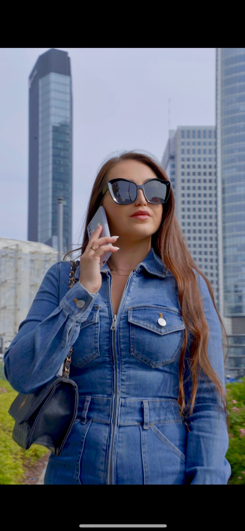 Kobieta w dżinsowym kombinezonie i dużych okularach przeciwsłonecznych rozmawia przez telefon na tle nowoczesnych biurowców.