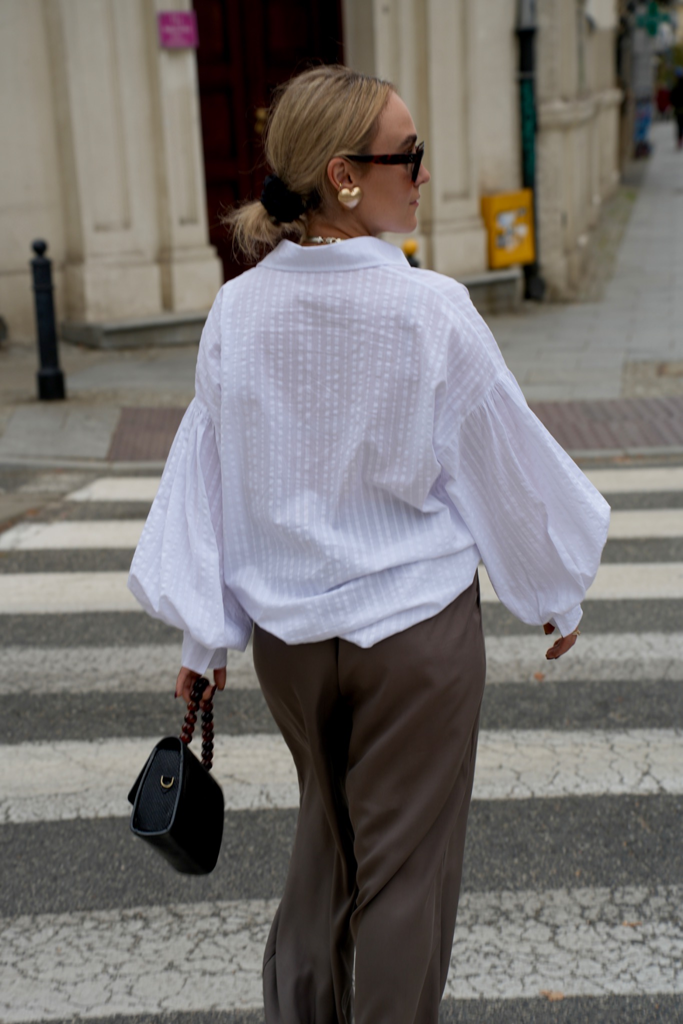 Kobieta w okularach przeciwsłonecznych, białej bluzce z bufiastymi rękawami i brązowych spodniach przechodzi przez przejście dla pieszych, trzymając czarną torebkę z brązowym uchwytem.