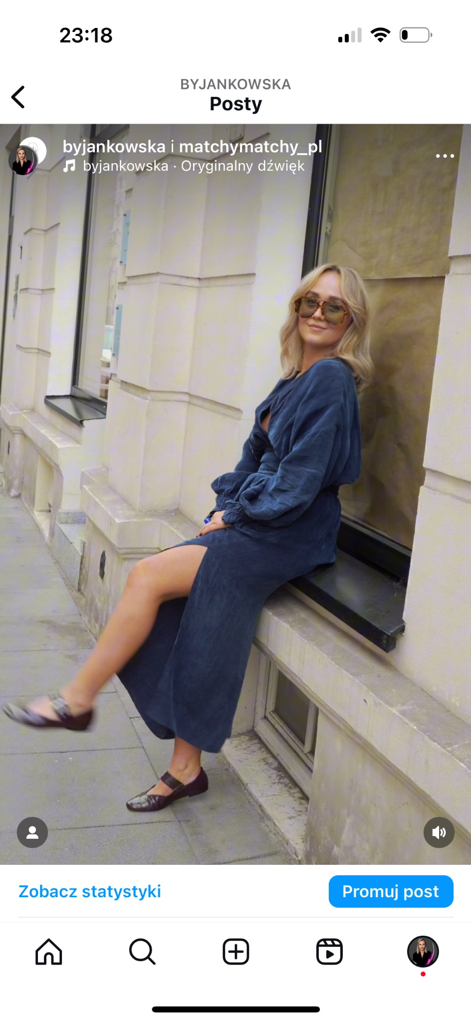 Kobieta w sukience w kolorze indygo i okularach przeciwsłonecznych siedzi na parapecie okna w kamienicy.