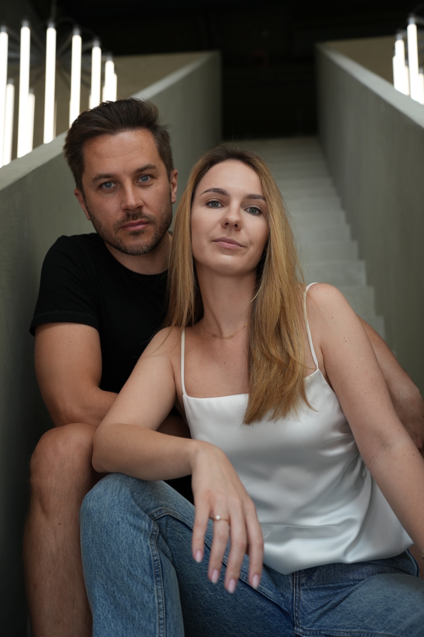 Portret pary siedzącej na betonowych schodach z nowoczesnym oświetleniem, kobieta w białym topie i dżinsach, mężczyzna w czarnym t-shircie.