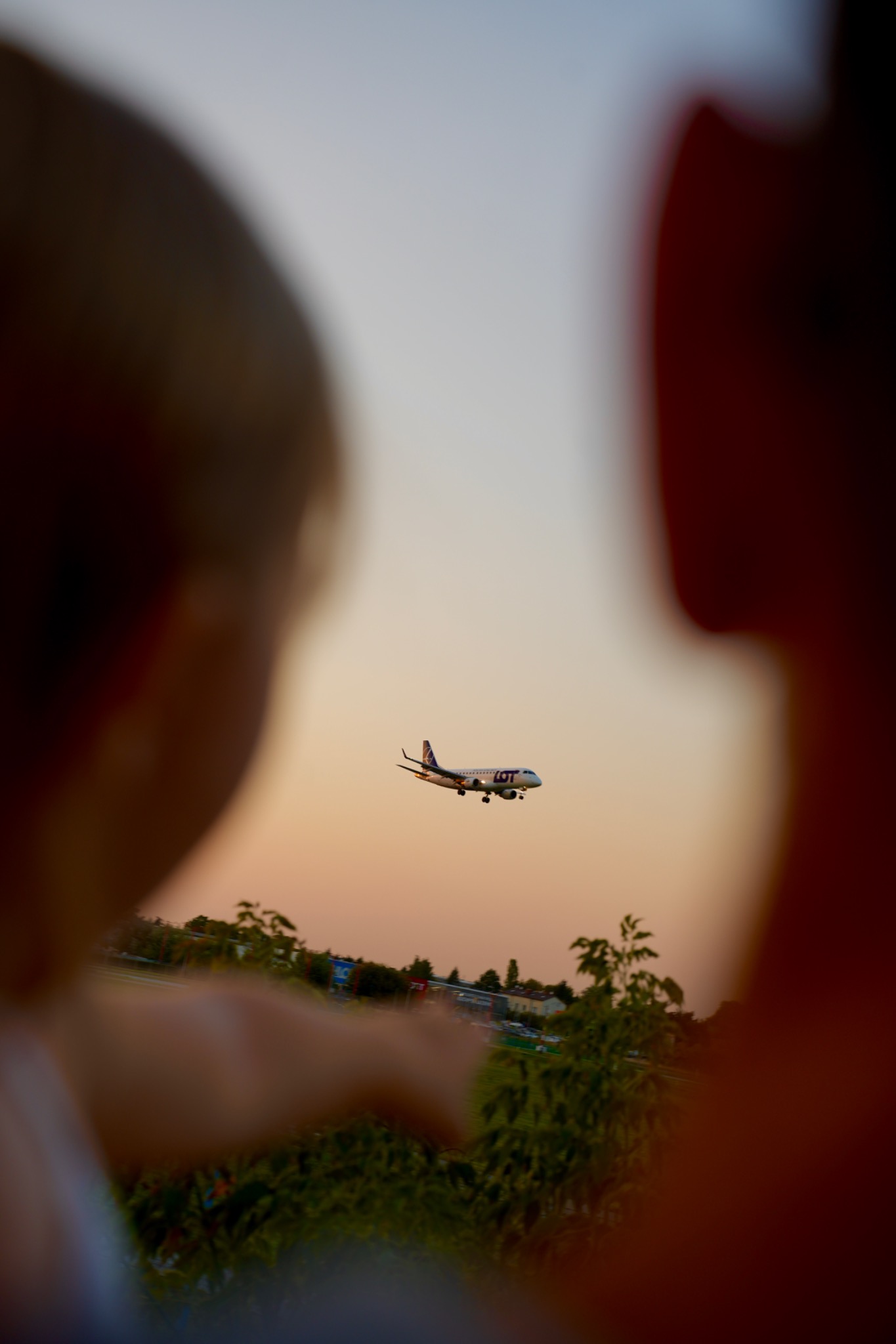 Samolot LOT w locie, widziany przez rozmyte sylwetki dziecka wskazującego palcem i dorosłego, na tle zachodzącego słońca i zieleni.