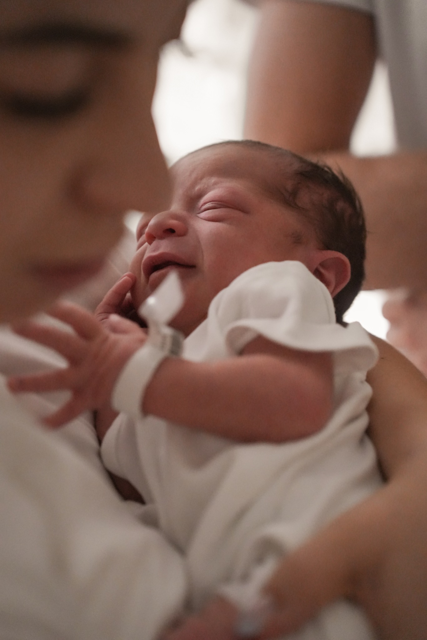 Noworodek z grymasem na twarzy, trzymany przez osobę w białej koszulce, z widoczną bransoletką identyfikacyjną na rączce dziecka.