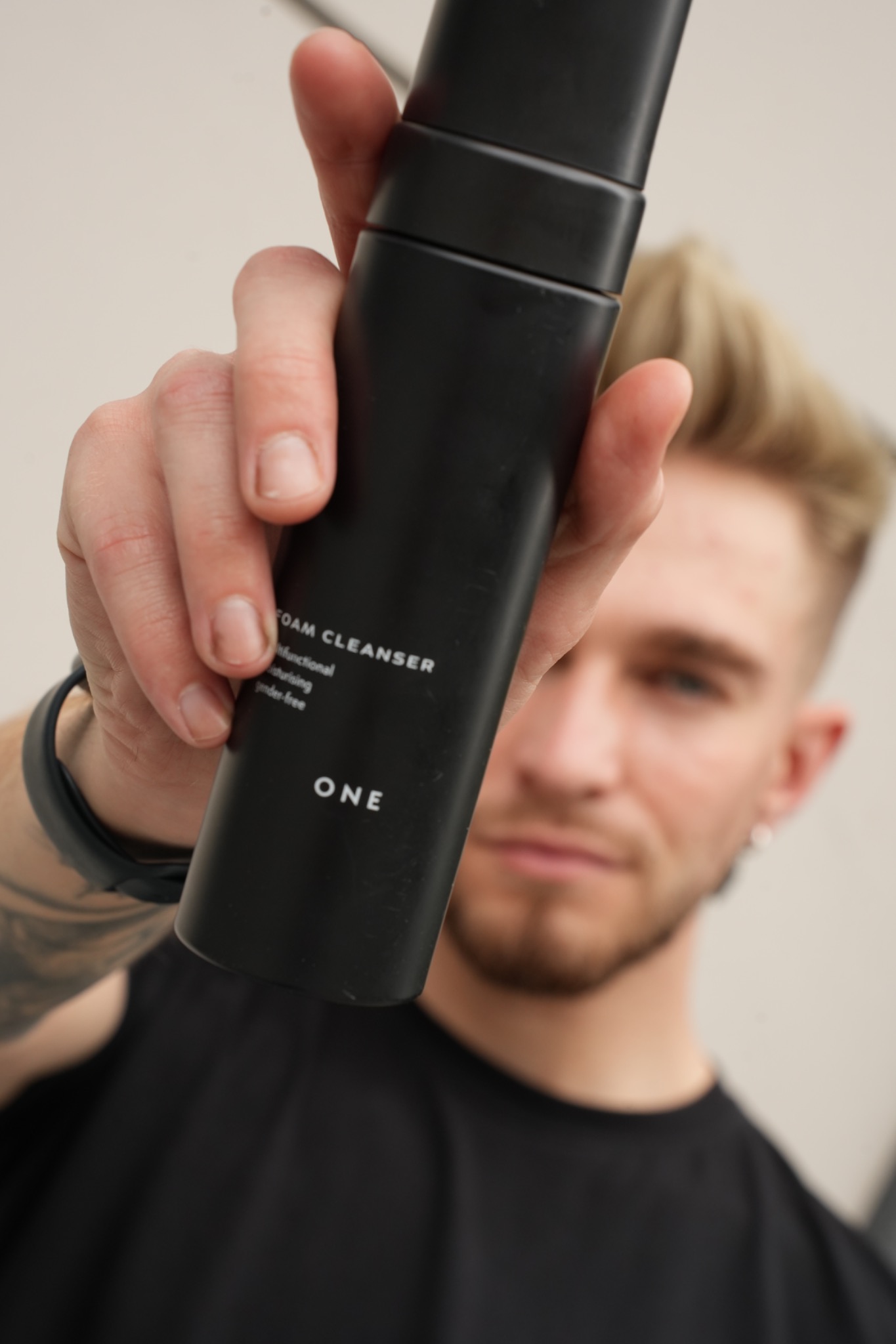 Mężczyzna trzyma w dłoni czarną butelkę pianki oczyszczającej do twarzy marki ONE, z tatuażem na przedramieniu i kolczykiem w uchu, na rozmytym tle ściany.