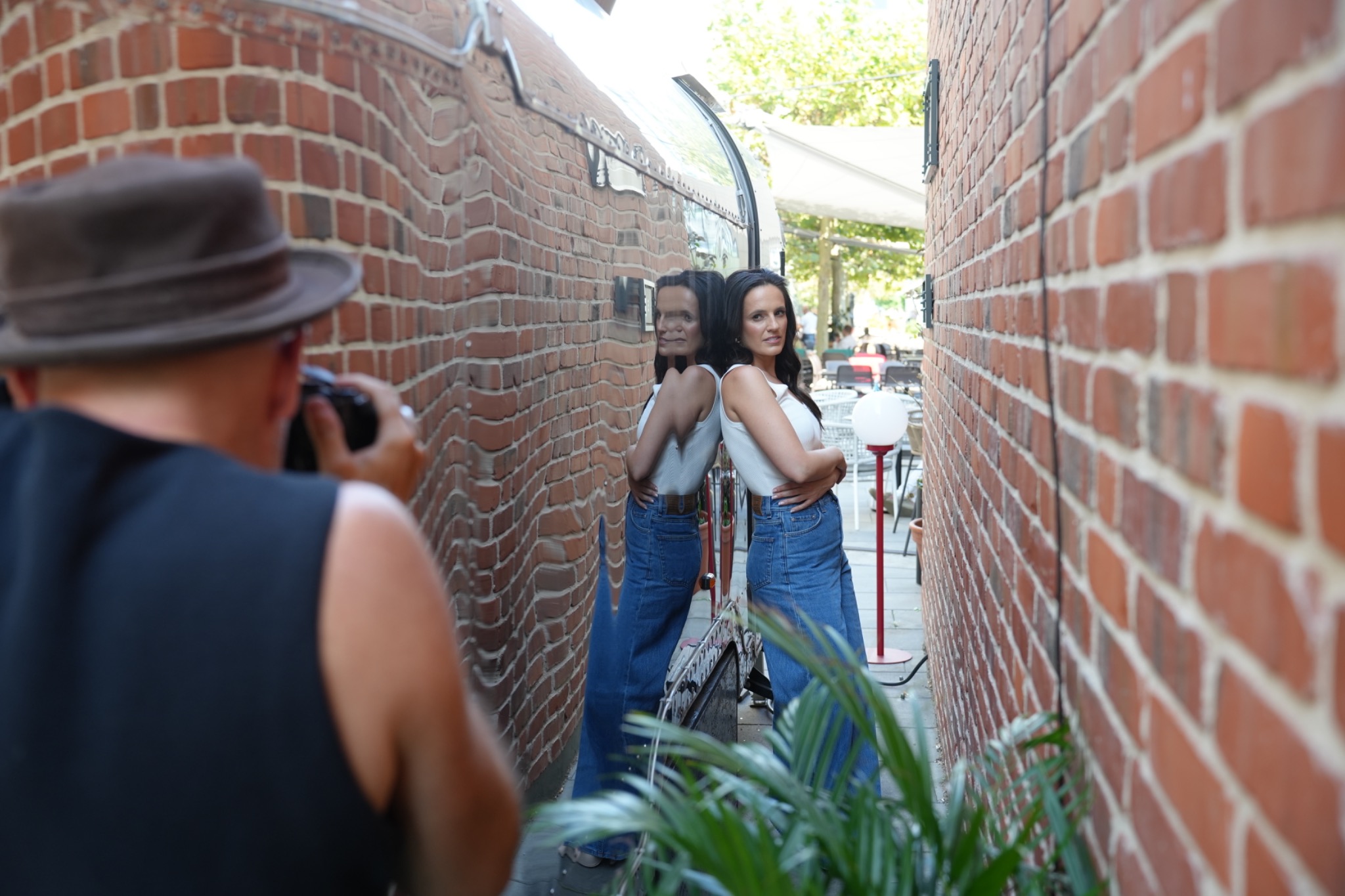 Kobieta w jeansach pozuje oparta o błyszczącą ścianę budynku, odbijającą ceglany mur, fotografowana przez osobę z aparatem w kapeluszu.