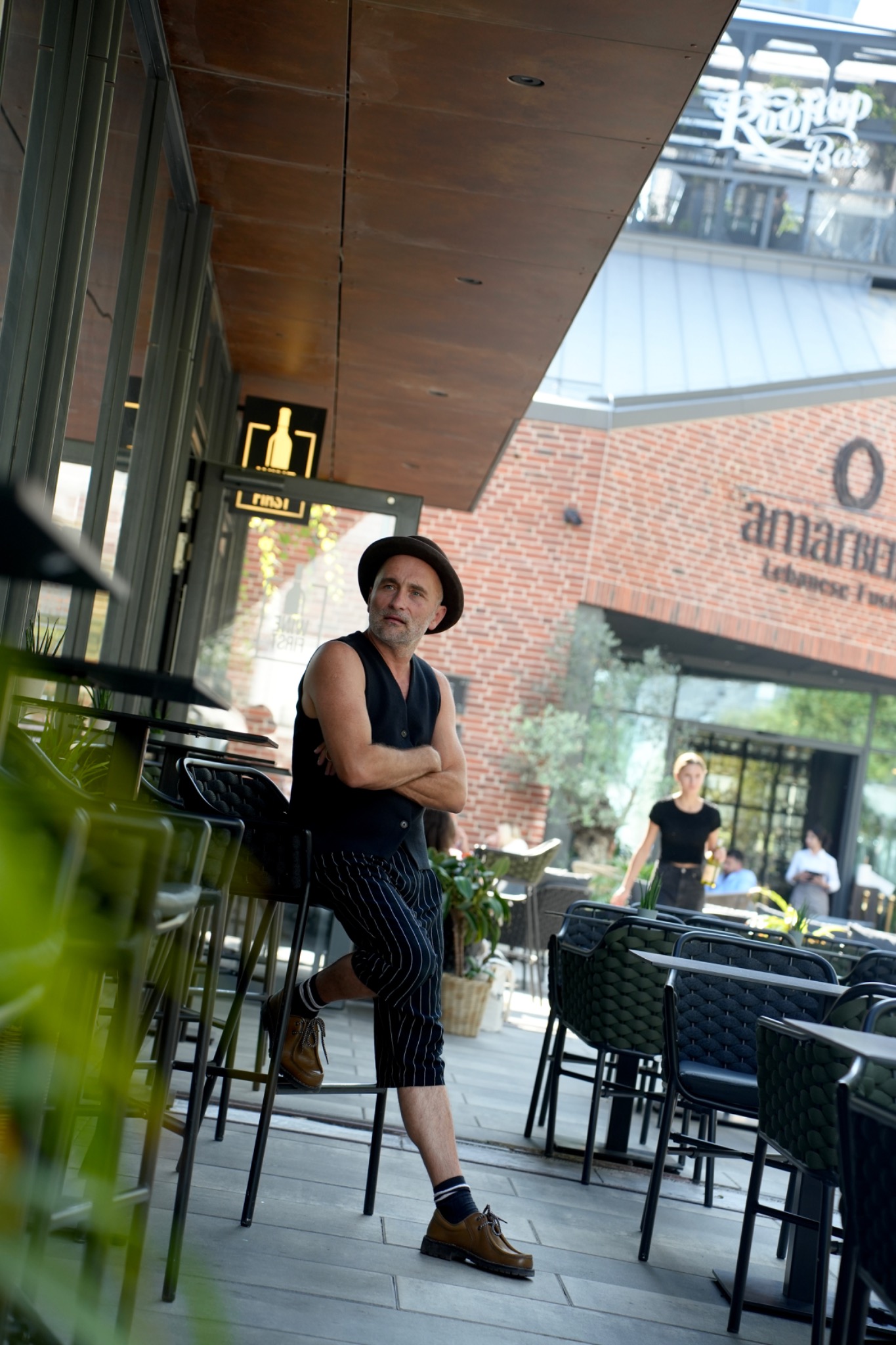 Mężczyzna w kapeluszu i kamizelce siedzi na wysokim krześle na zewnątrz restauracji z napisem 'amarBER' i 'Rooftop Bar' w tle.