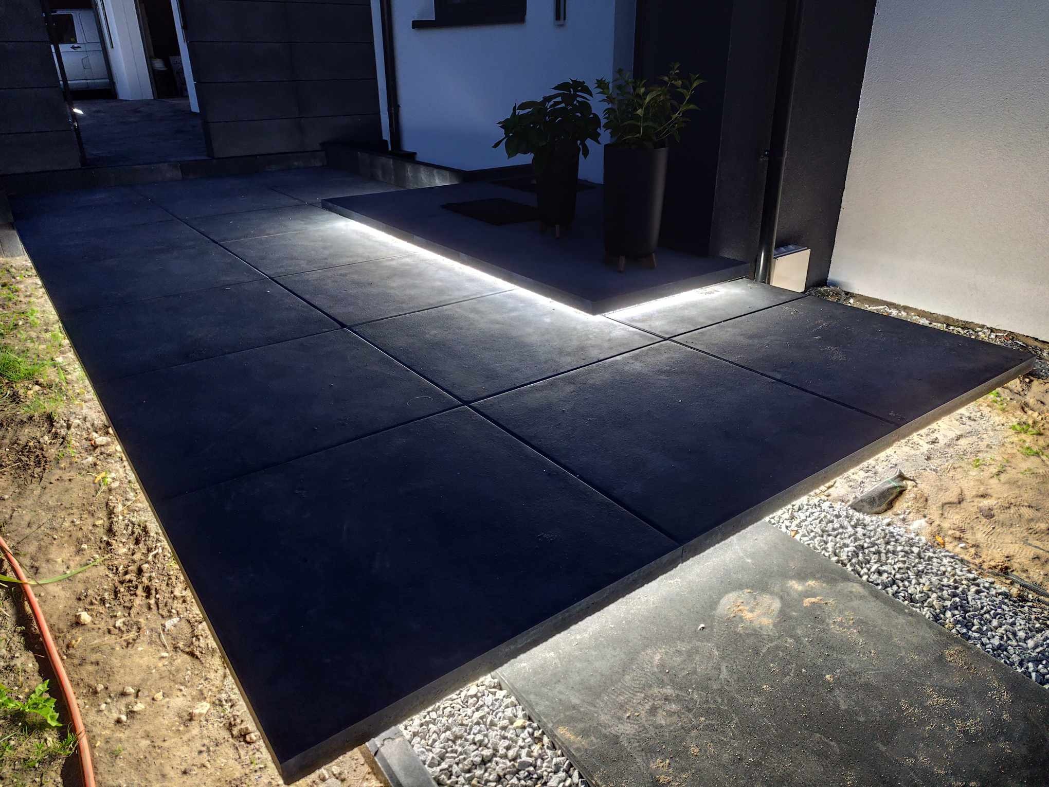 Nowoczesna aranżacja wejścia do domu z podświetlanymi płytami chodnikowymi i donicami z roślinami, oświetlenie LED wzdłuż krawędzi ścieżki.