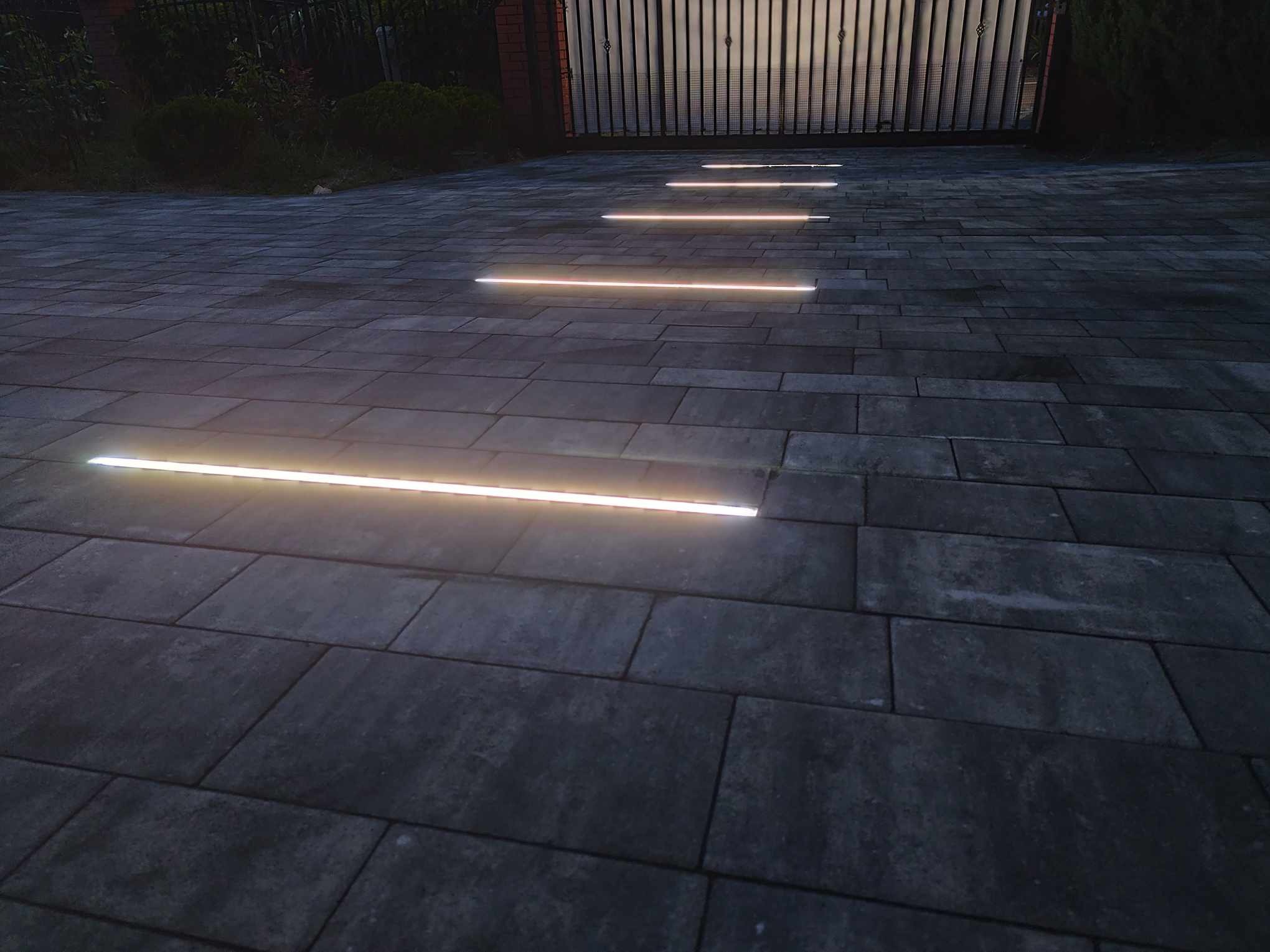 Świecące, poziome listwy LED wkomponowane w szarą kostkę brukową podjazdu, prowadzące do bramy wjazdowej. Nastrojowe oświetlenie zewnętrzne.