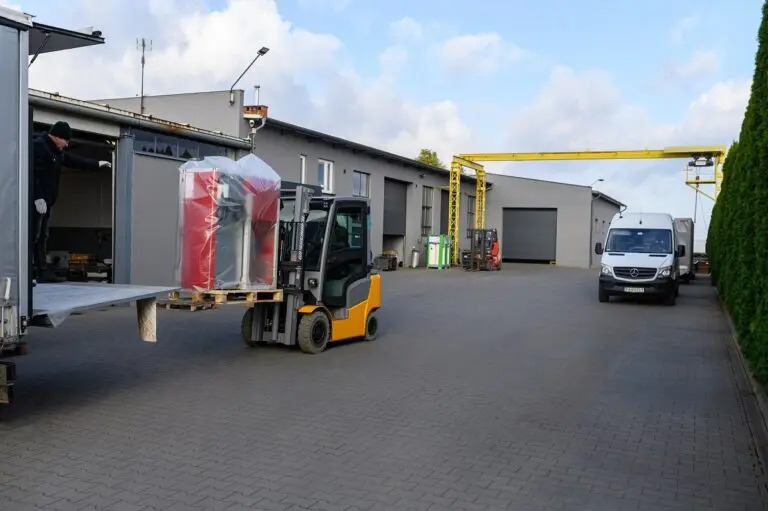 Wózek widłowy transportuje dwa piece CO na palecie z ciężarówki, pracownik w czapce kieruje załadunkiem, w tle budynek magazynowy i zaparkowane pojazdy.