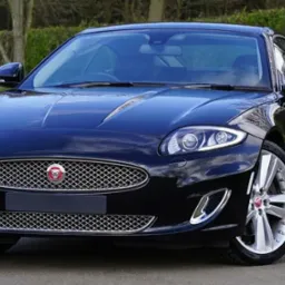 Czarny samochód Jaguar XK z aluminiowymi felgami, widok z przodu na tle zieleni.