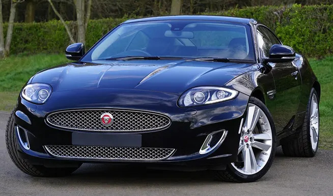 Czarny samochód Jaguar XK z aluminiowymi felgami, widok z przodu na tle zieleni.