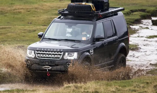 Czarny Land Rover Discovery z bagażnikiem dachowym i kołem zapasowym pokonuje błotnisty teren, widoczne chlapiące błoto i hak holowniczy.