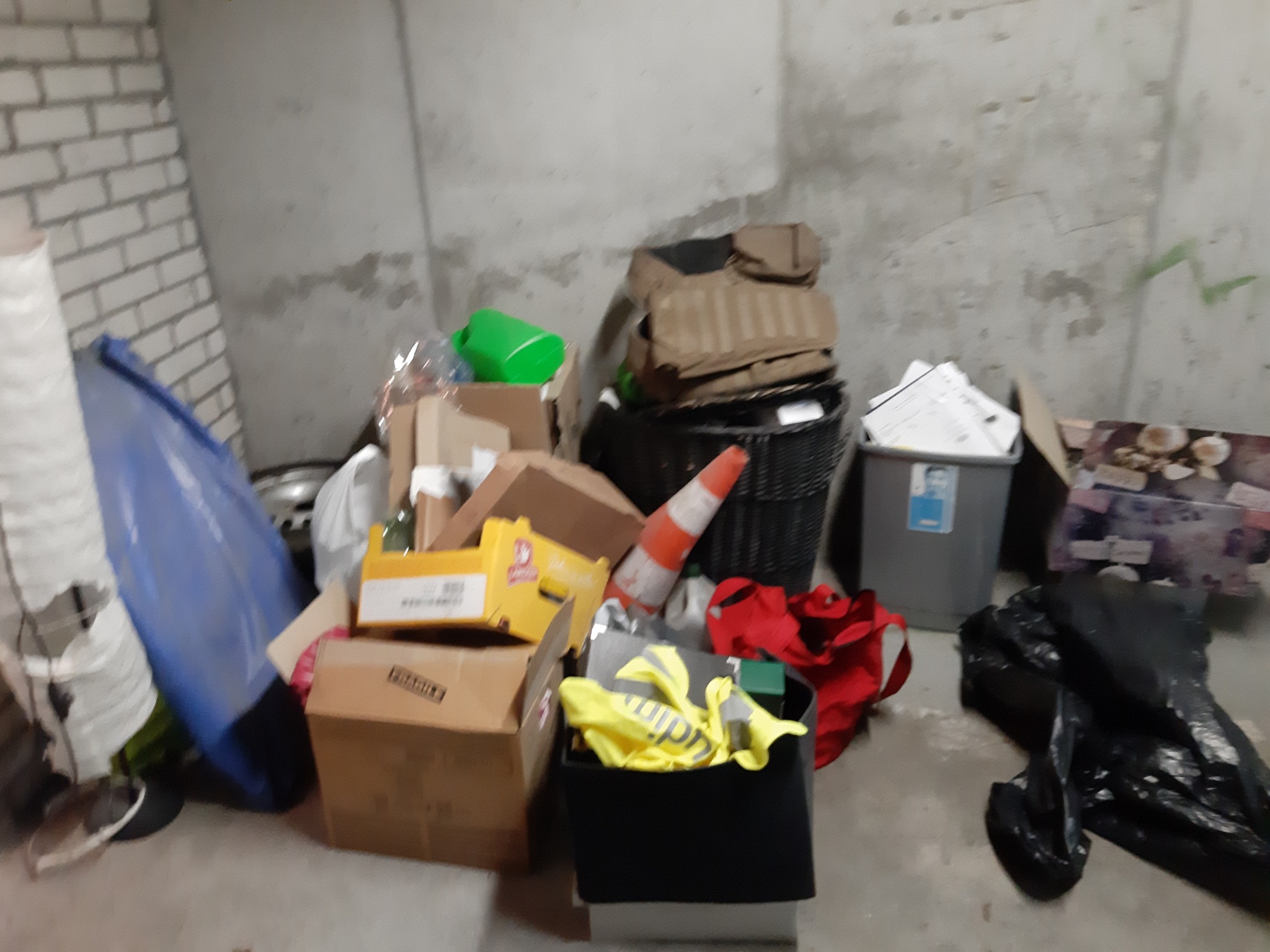 Sterta różnorodnych przedmiotów i odpadów, w tym kartony, plastikowe pojemniki, odzież robocza i worki na śmieci, zgromadzona w piwnicy z surowymi ścianami z cegły i betonu.