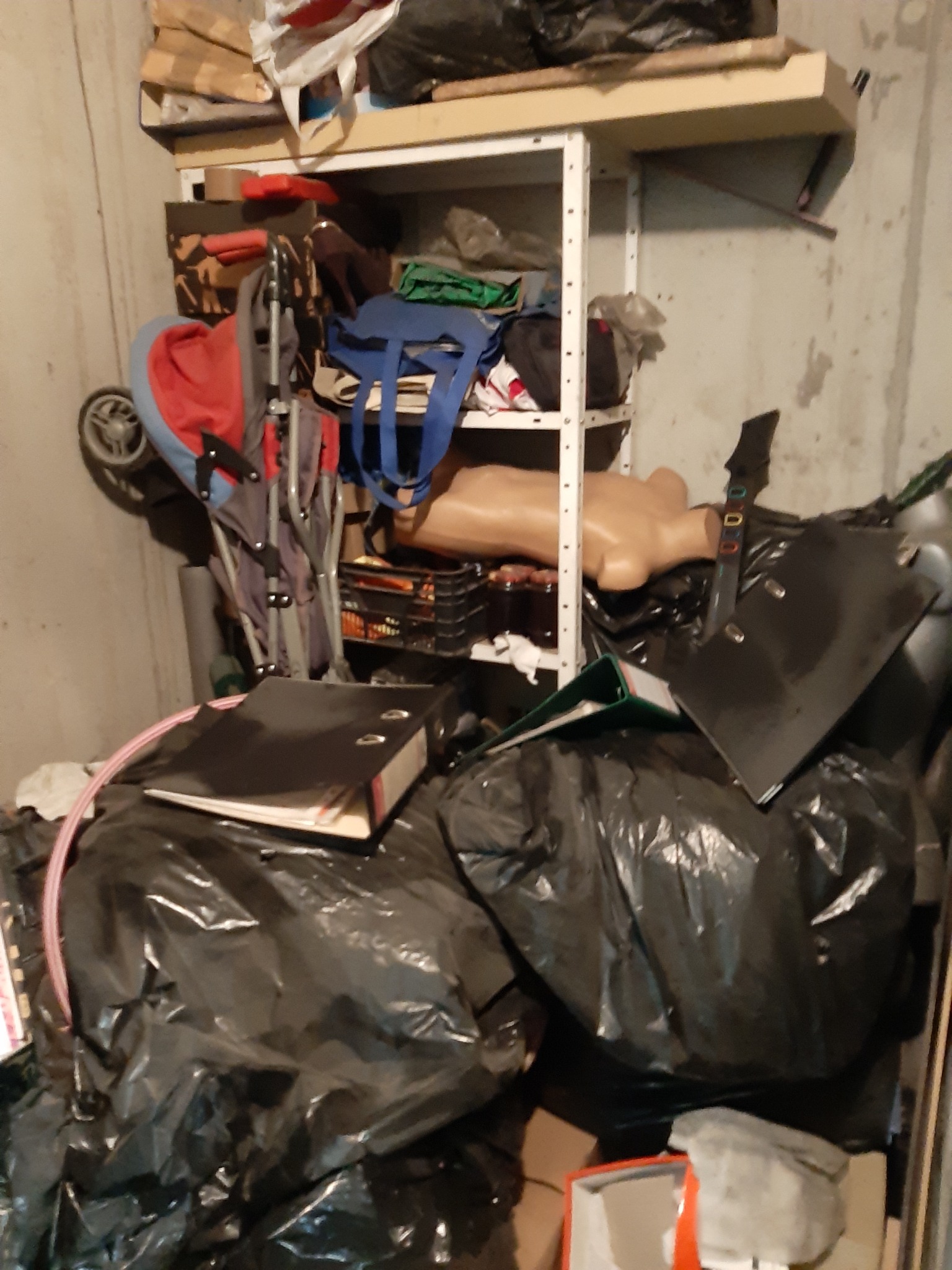 Zagracone pomieszczenie z metalowym regałem, na którym znajdują się różne przedmioty, w tym manekin, wózek dziecięcy i torby z zawartością.