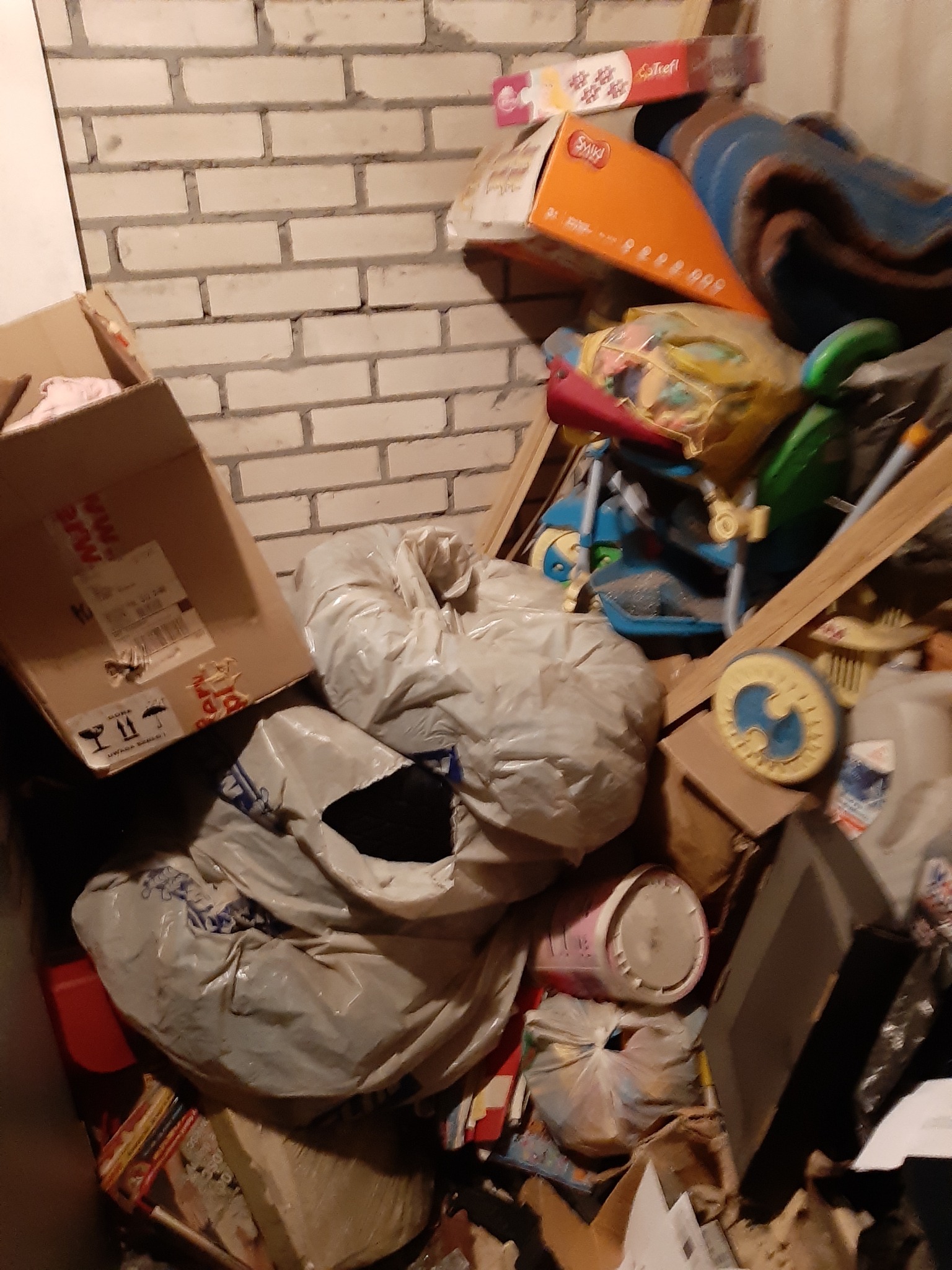 Stos kartonów, worków z zawartością, zabawek i innych przedmiotów przy ceglanej ścianie, w pomieszczeniu słabo oświetlonym.