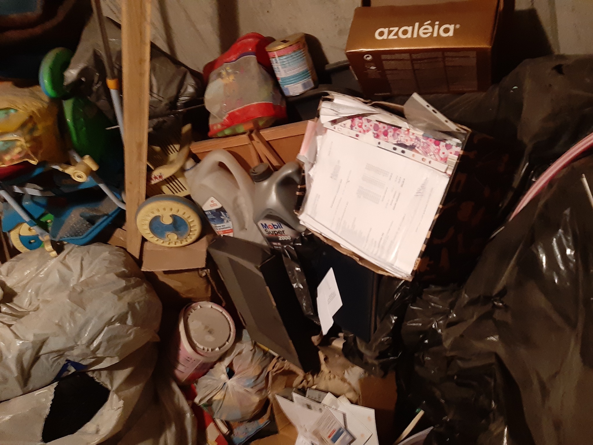 Stos różnych przedmiotów, w tym zabawki, puszki, kartony, butelki po oleju samochodowym i worki na śmieci, zgromadzonych w słabo oświetlonym pomieszczeniu.