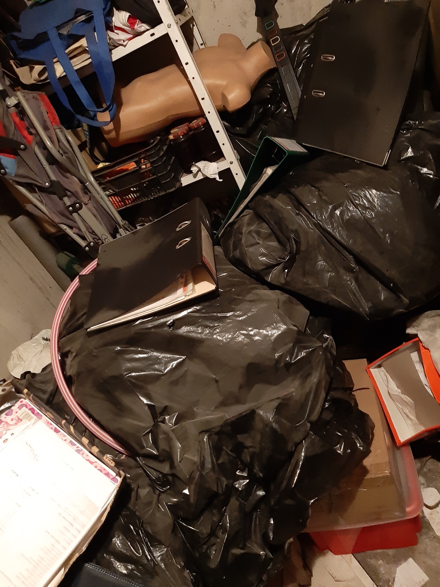 Wnętrze zagraconego pomieszczenia: czarne worki na śmieci, segregator, manekin bez głowy na metalowym regale, pudła i inne przedmioty.