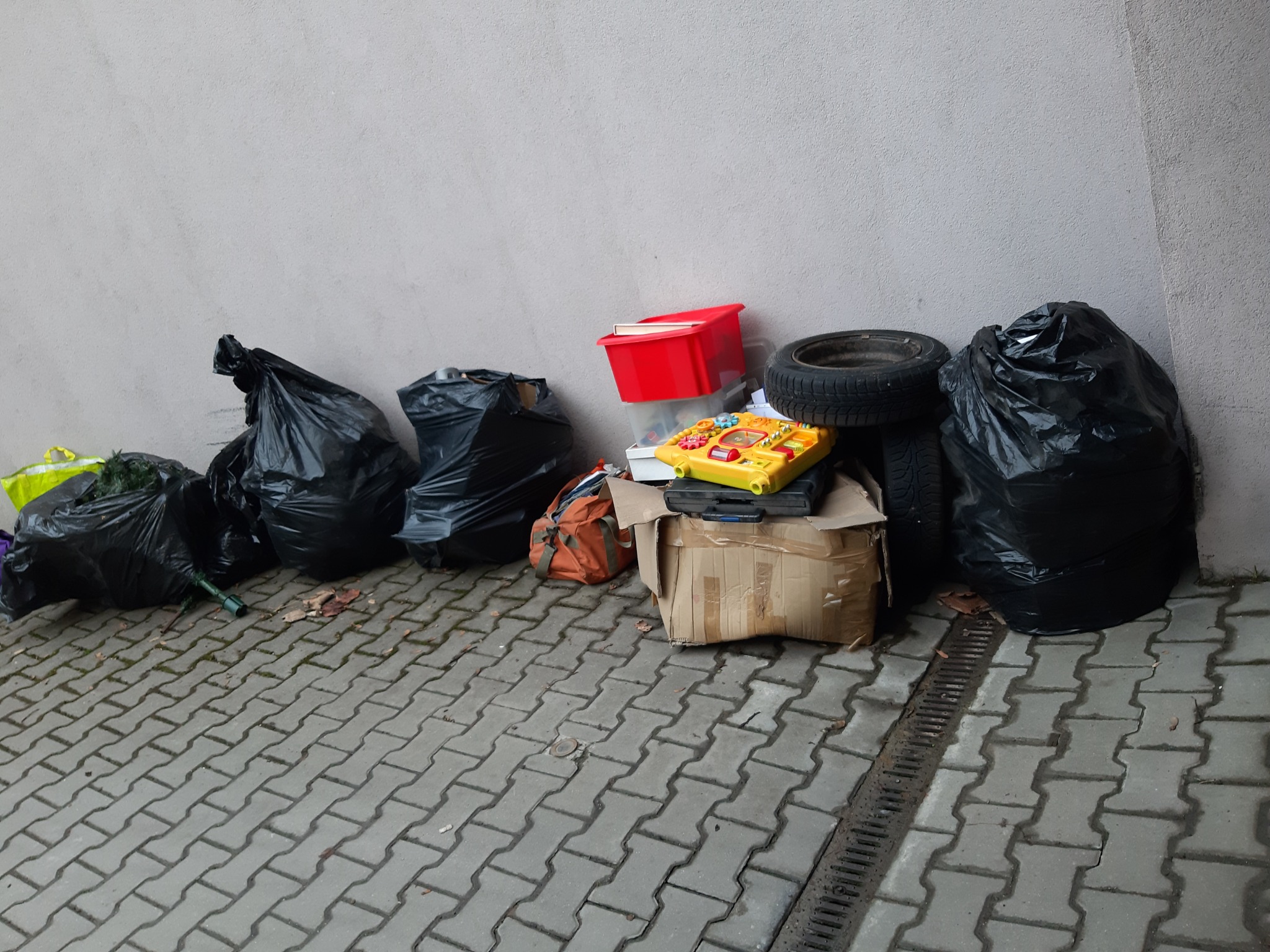 Stos czarnych worków na śmieci, starych opon, pudeł kartonowych, czerwonego pojemnika i zabawek ustawionych na chodniku z kostki brukowej przy szarej ścianie.