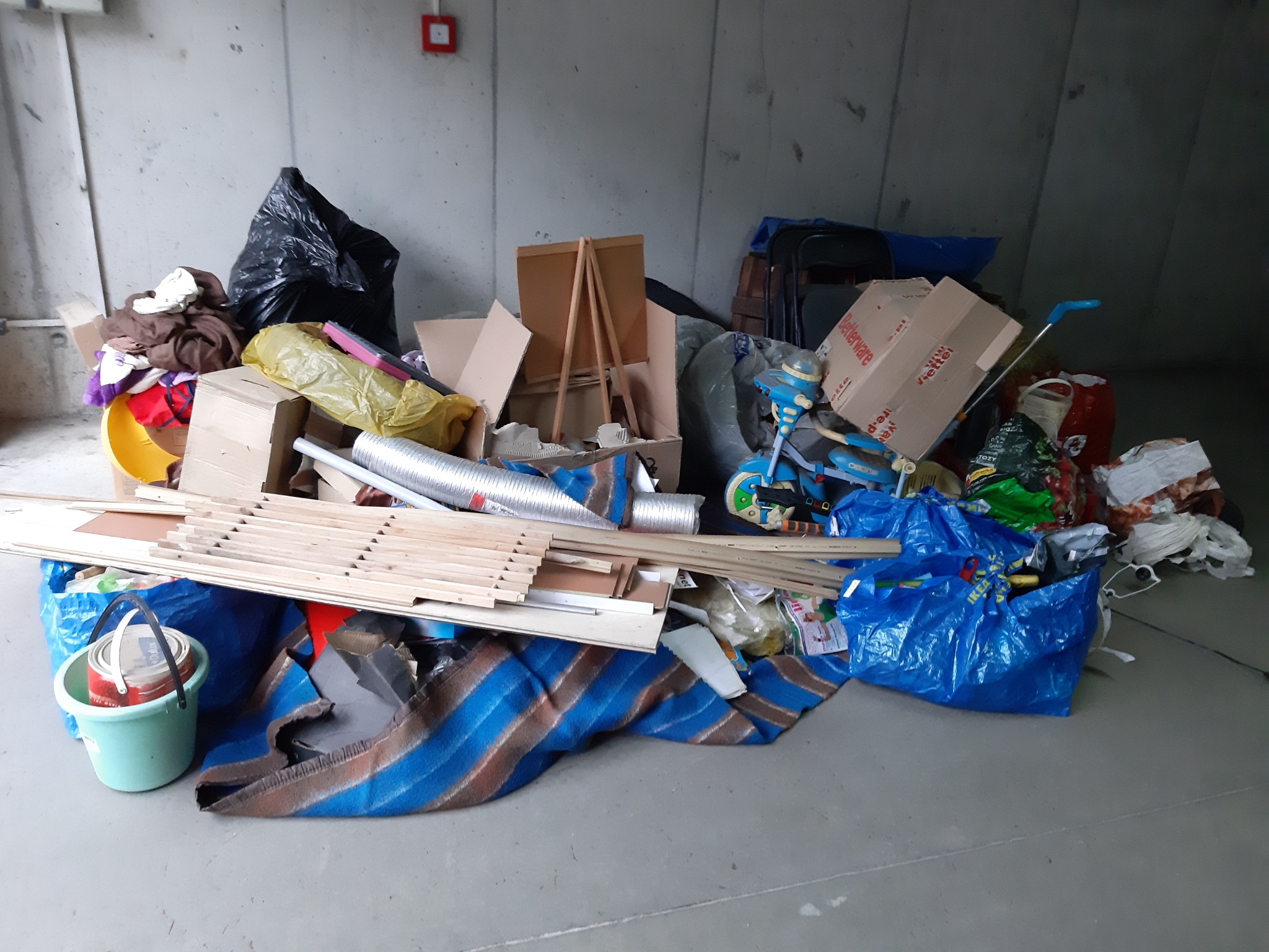 Sterta różnorodnych przedmiotów i odpadów, w tym meble do samodzielnego montażu, zabawki dziecięce, tekstylia, puszka z farbą i worki na śmieci, zgromadzona na betonowej podłodze w pomieszczeniu...