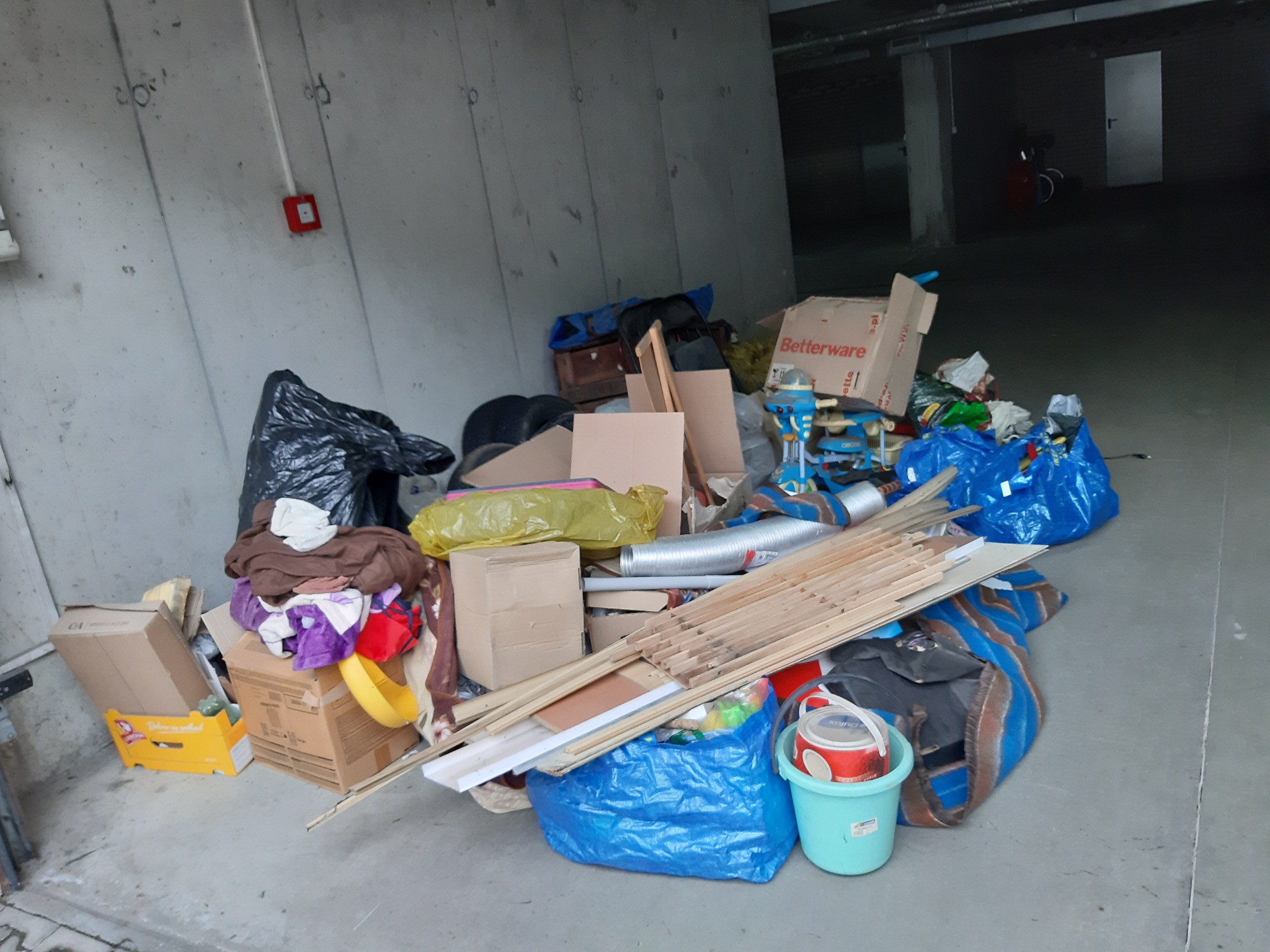 Sterta różnorodnych odpadów, w tym kartony, worki na śmieci, resztki materiałów budowlanych i zużyte przedmioty gospodarstwa domowego, nagromadzona w rogu garażu lub piwnicy, z widocznym alarmem...