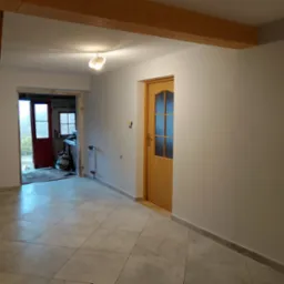 Wnętrze domu w trakcie odświeżania: pomalowane ściany na jasny kolor, nowa podłoga z płytek, drewniane belki stropowe, drzwi wewnętrzne z przeszkleniami i widok na pomieszczenie gospodarcze...