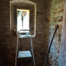 Usługi remontowo-budowlane - Wnętrze pomieszczenia z surowymi ścianami z cegły, drabina aluminiowa oparta o ścianę, okno z narzędziami i pudełkiem, widoczne ślady prac remontowych.
