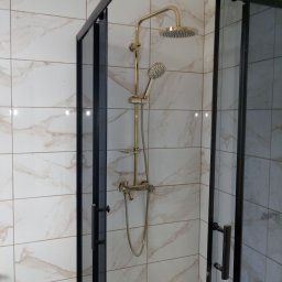 Usługi remontowo-budowlane - Kabina prysznicowa z czarną ramą i złotą armaturą na tle marmurowych płytek.
