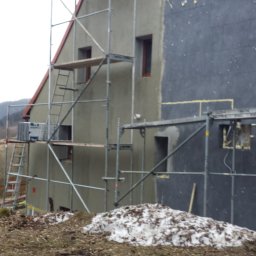 Usługi remontowo-budowlane - Dom w trakcie prac elewacyjnych: jedna ściana ocieplona i pokryta szarą płytą, druga w trakcie szpachlowania, rusztowania ustawione przy ścianach, w tle las.