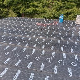 Usługi remontowo-budowlane - Widok z góry na fragment dachu pokrytego czarną papą z nadrukiem IzoBud, w tle zielone drzewa iglaste i niebieska butla gazowa z czerwoną rurką.