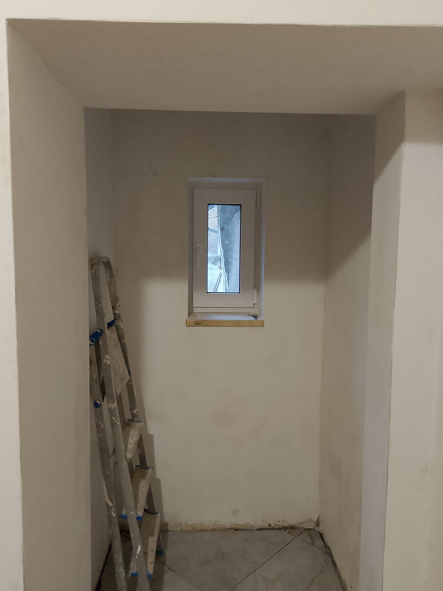 Wnęka z małym oknem w trakcie prac wykończeniowych; widoczna drabina malarska oparta o ścianę, surowe ściany przygotowane do malowania, szare płytki podłogowe.