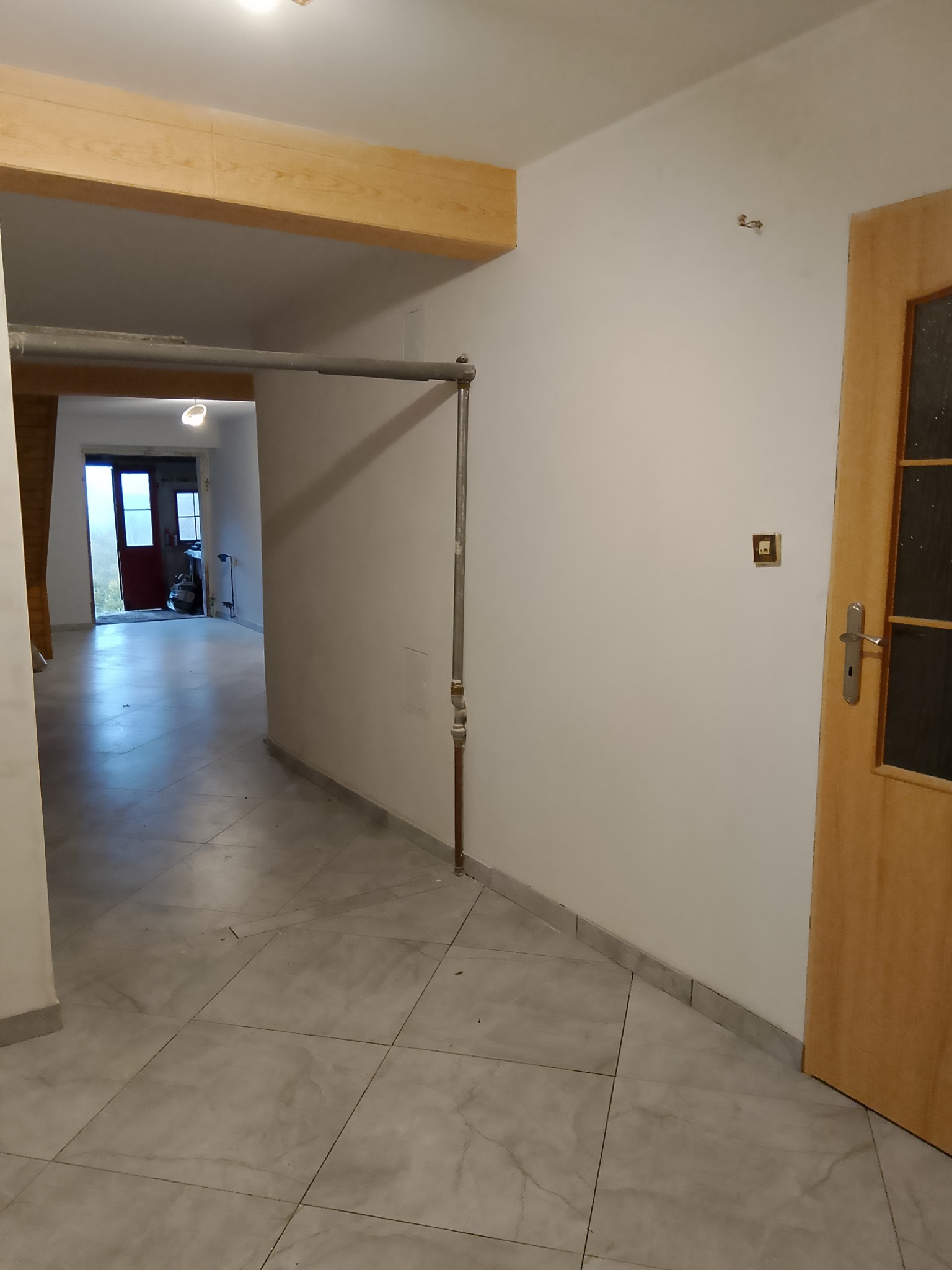 Wnętrze korytarza w trakcie remontu: szare płytki podłogowe, białe ściany z widoczną instalacją elektryczną, drewniana belka stropowa i ościeżnica drzwiowa.