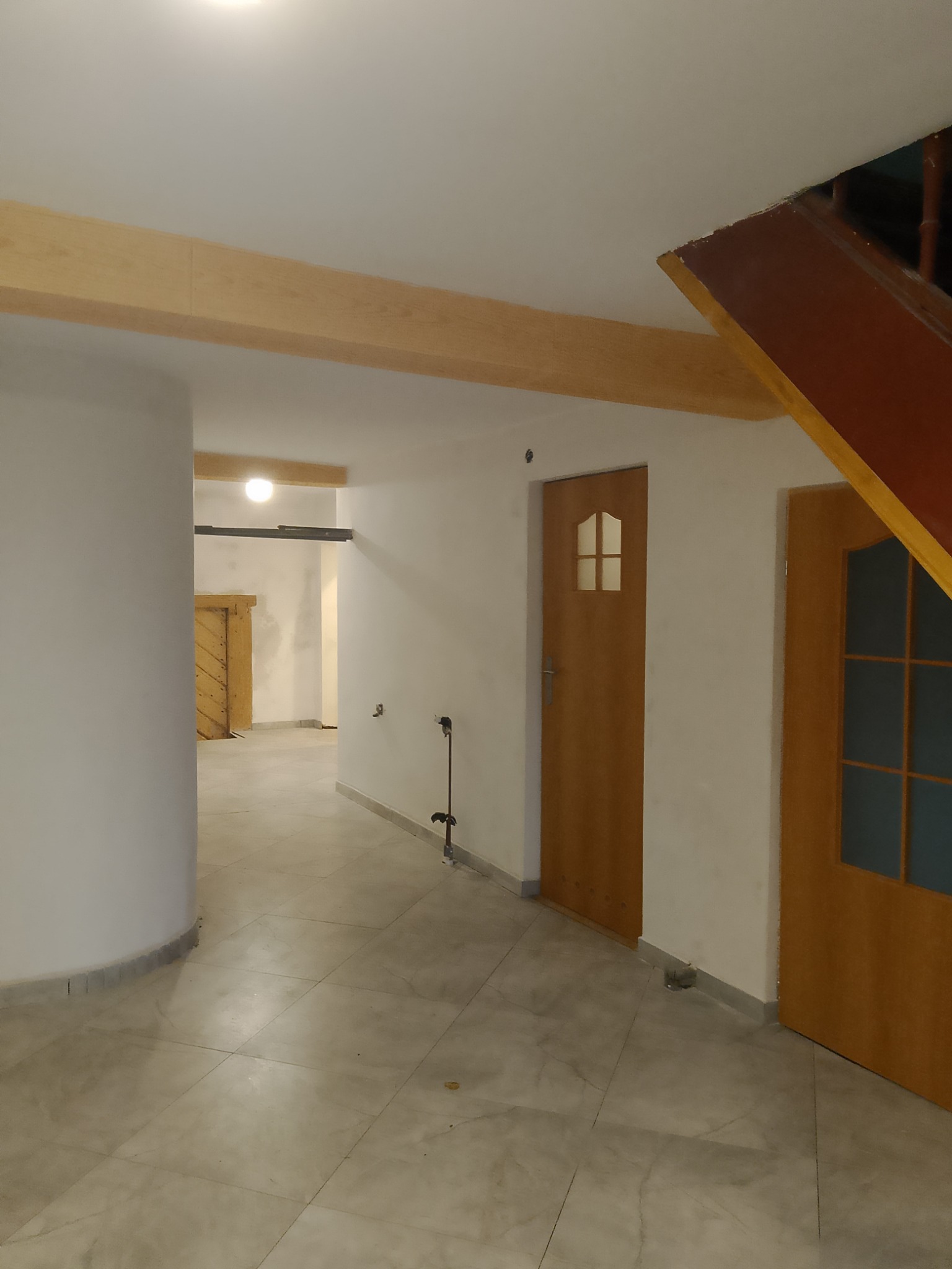 Korytarz w domu z drewnianymi belkami stropowymi, dwiema drewnianymi drzwiami i wykończoną podłogą z płytek imitujących marmur.