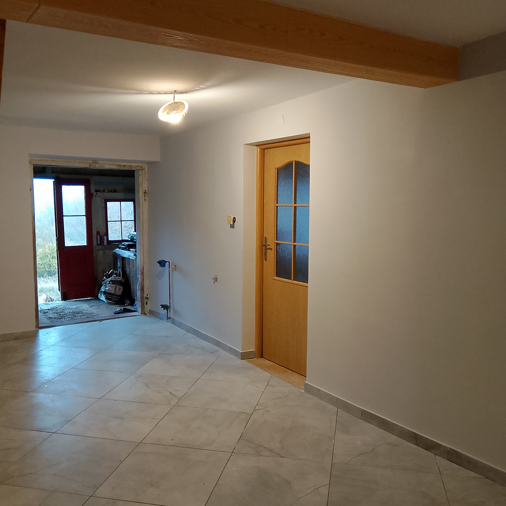 Wnętrze domu w trakcie odświeżania: pomalowane ściany na jasny kolor, nowa podłoga z płytek, drewniane belki stropowe, drzwi wewnętrzne z przeszkleniami i widok na pomieszczenie gospodarcze...