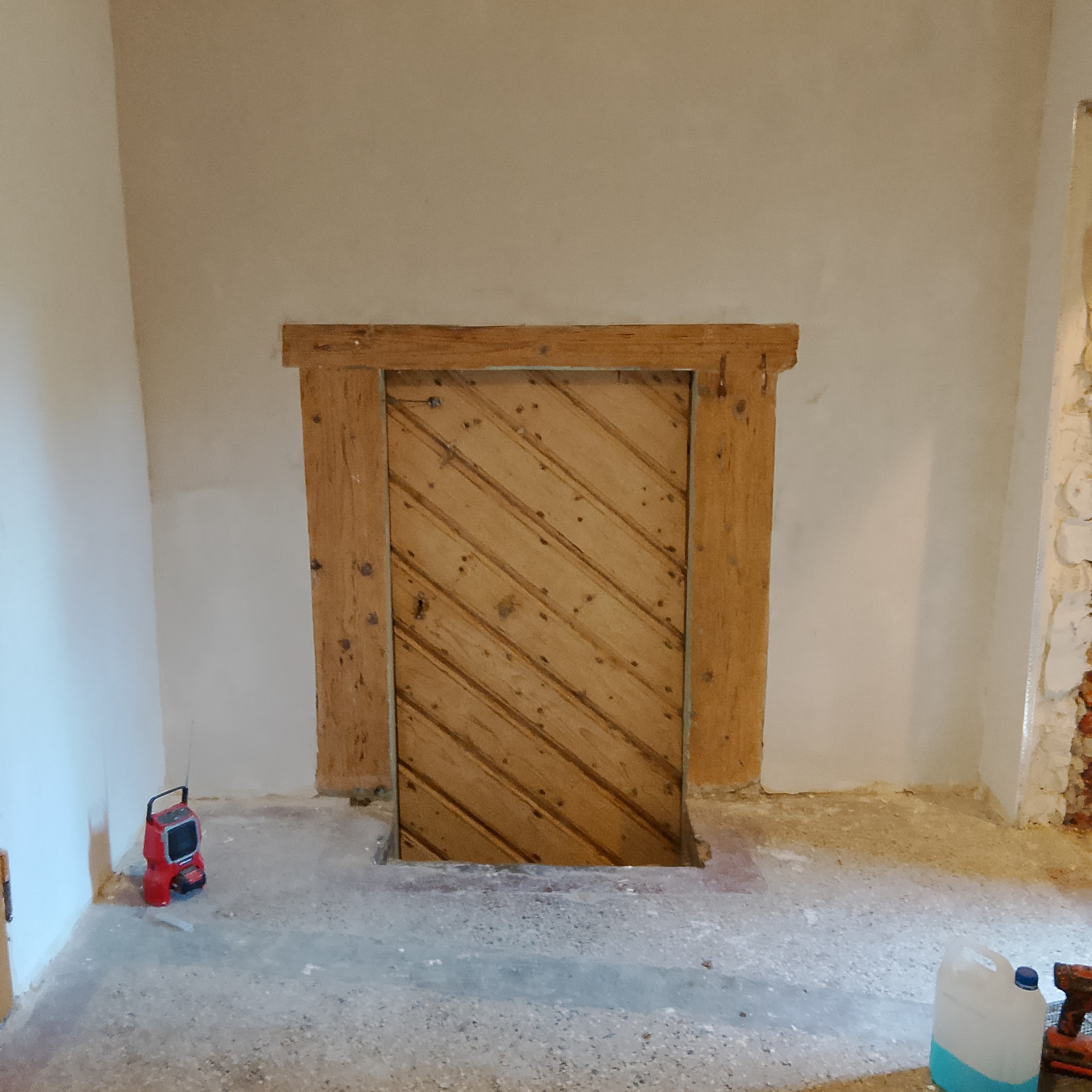 Drewniane drzwi z ukośnymi deskami w surowej ościeżnicy, w trakcie prac wykończeniowych; widoczny fragment ściany z odsłoniętą cegłą i narzędzia na podłodze.