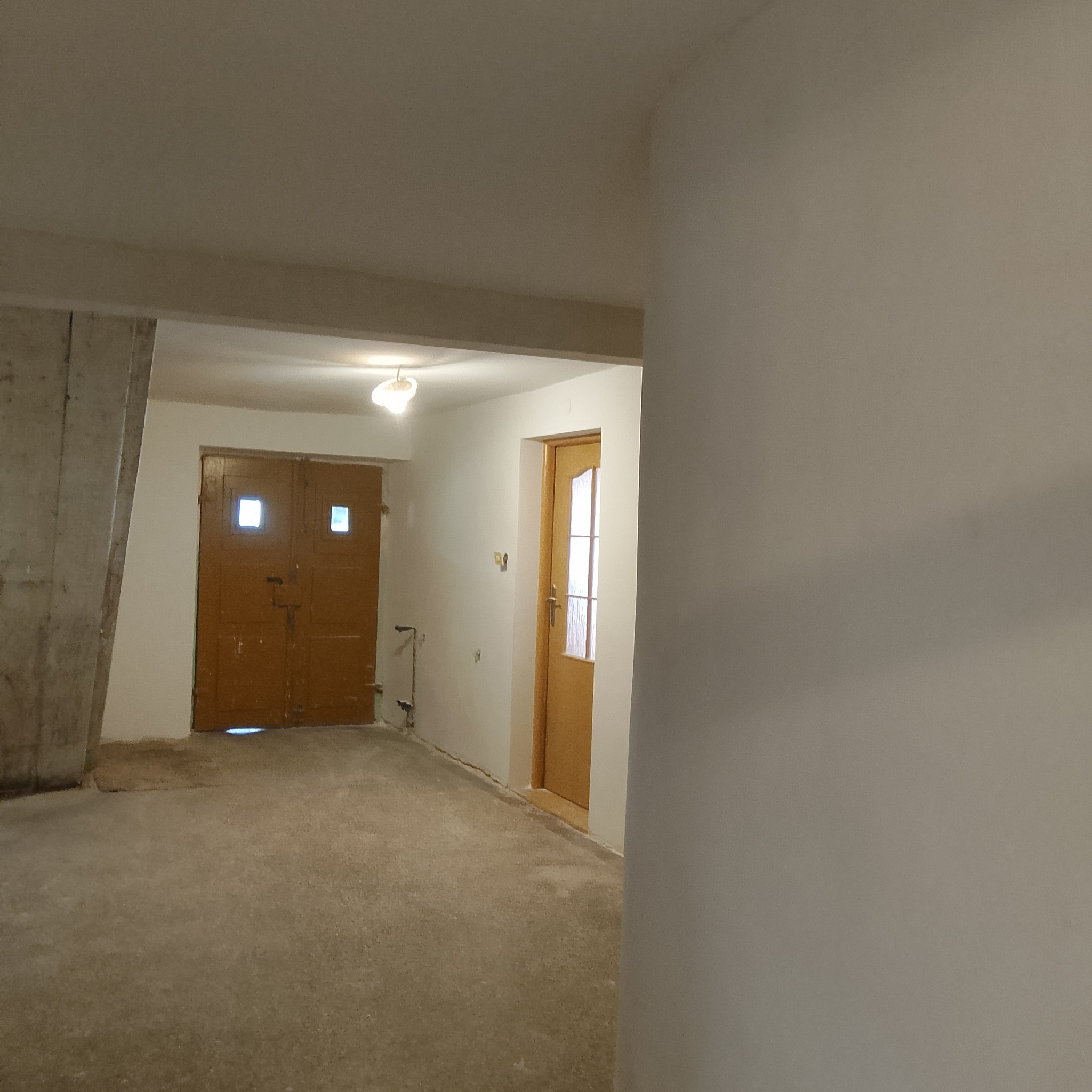 Wnętrze w trakcie remontu: świeżo pomalowane ściany na biało, widoczne dwie drewniane drzwi, surowy betonowy filar i niedoskonałości podłogi.