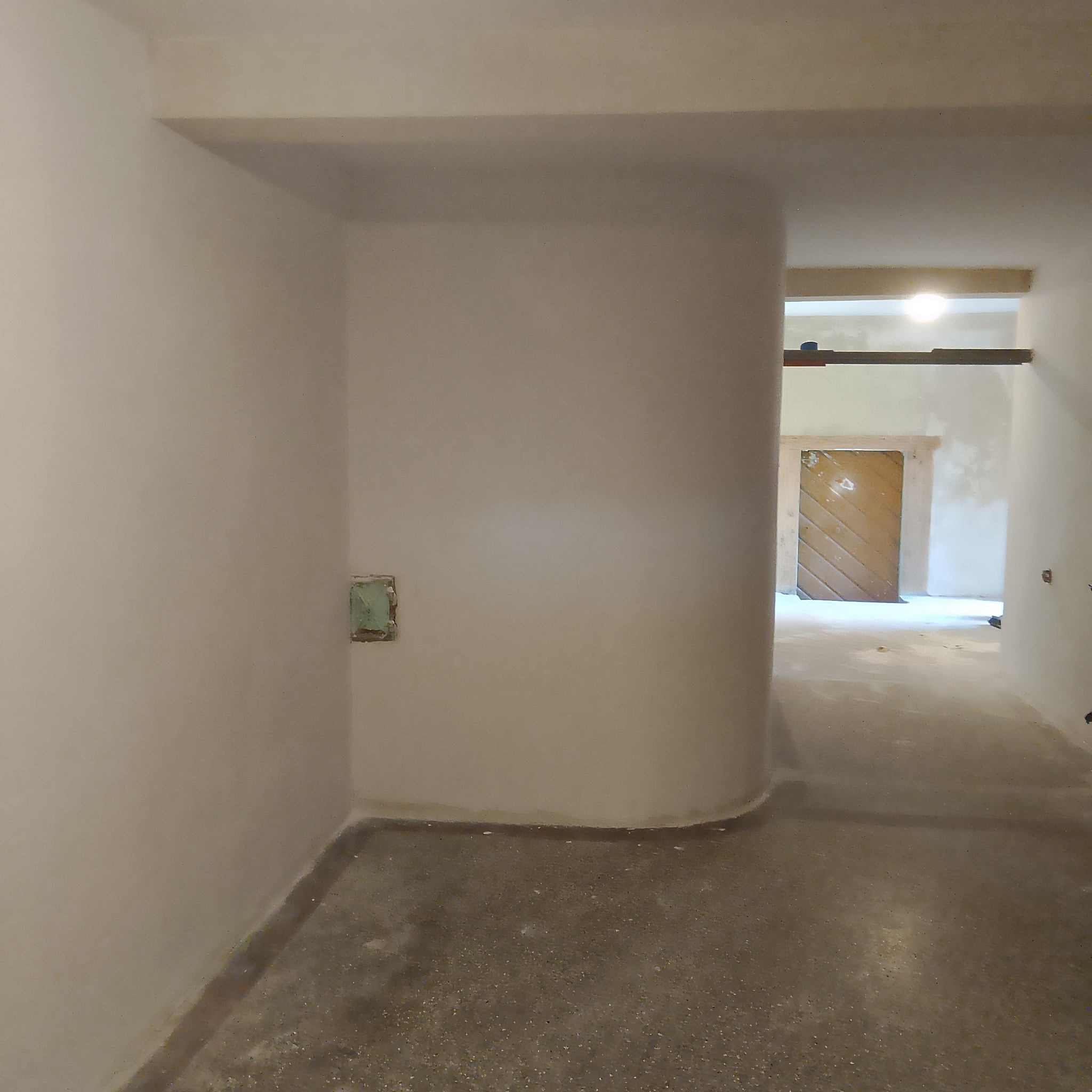 Wnętrze w trakcie remontu: surowe ściany w jasnym odcieniu, betonowa podłoga, widoczne ślady prac budowlanych i fragment drewnianych belek stropowych.
