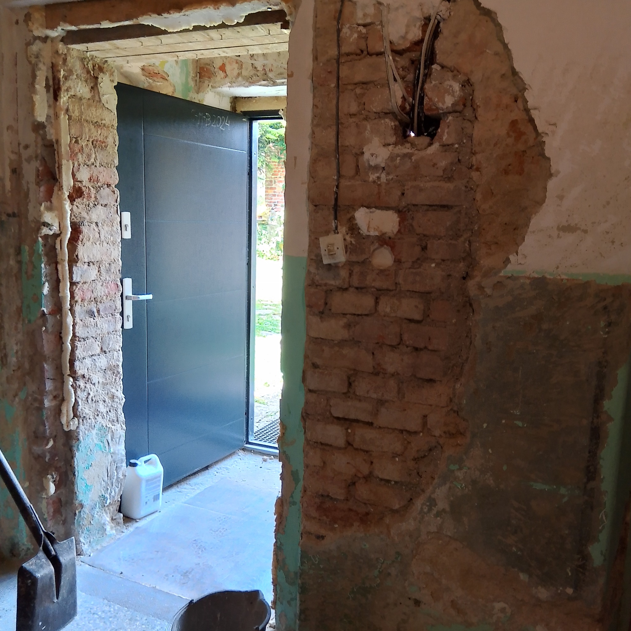 Otwór drzwiowy w trakcie remontu: widoczne fragmenty cegieł, uszkodzony tynk, wystające przewody elektryczne, częściowo zamontowane nowe drzwi wejściowe z widoczną datą na skrzydle, pojemnik...
