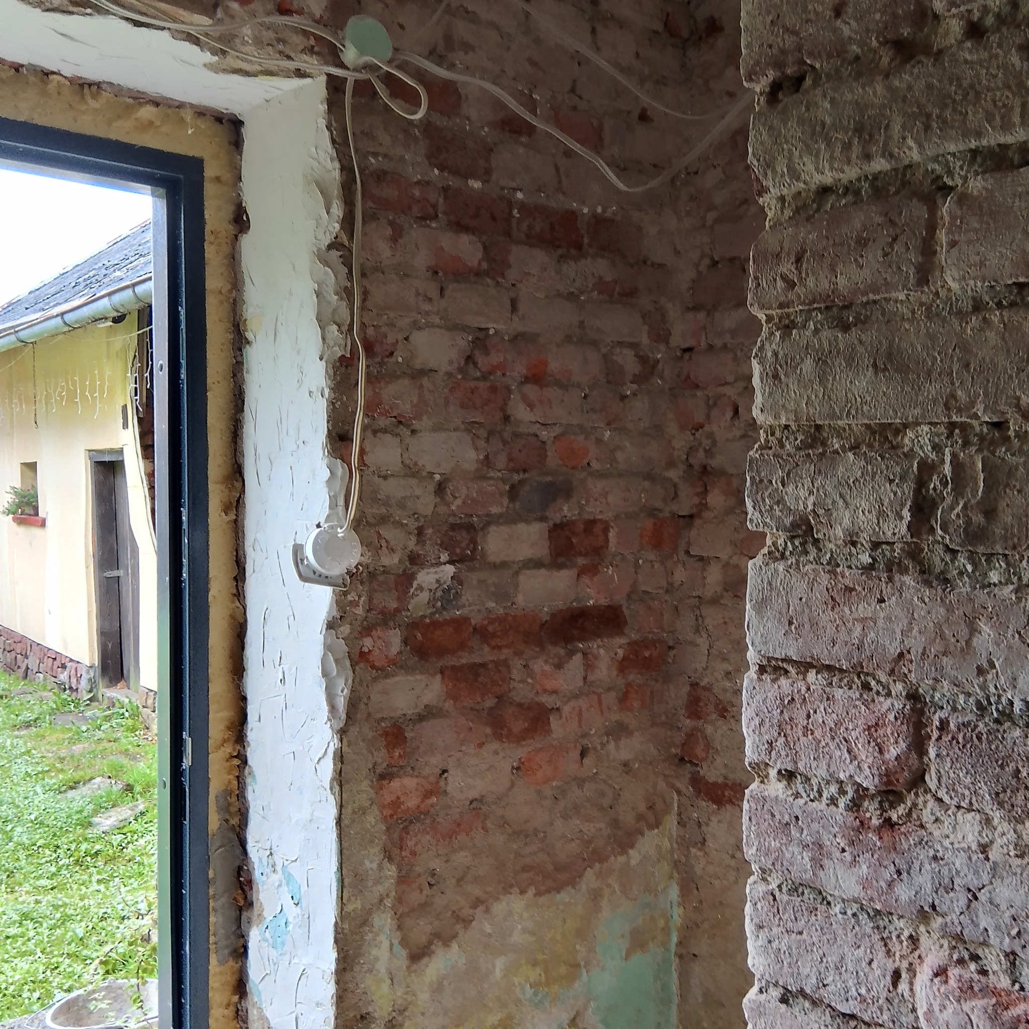 Wnętrze w trakcie remontu: odsłonięta cegła, przygotowywana do dalszych prac, z widoczną instalacją elektryczną i oknem z widokiem na zewnątrz.
