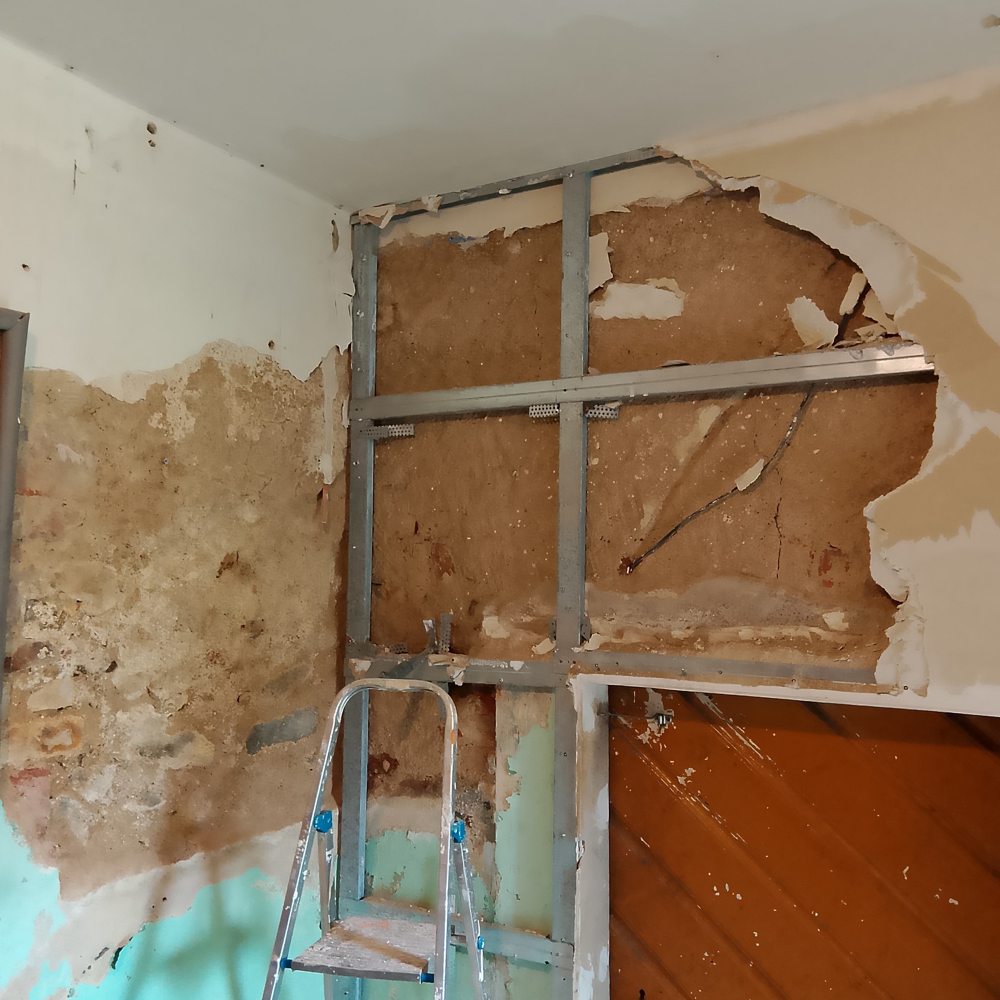Demontaż ściany działowej: odsłonięta konstrukcja metalowa, fragmenty starego tynku i instalacji elektrycznej, drabina oparta o ścianę.