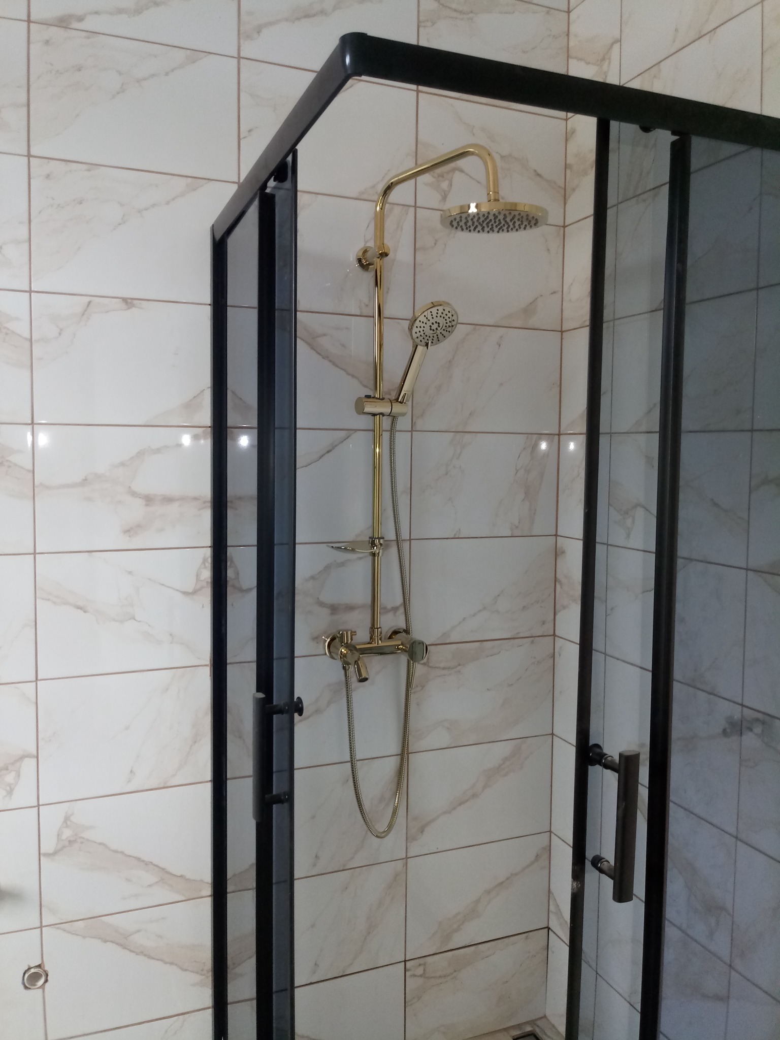 Kabina prysznicowa z czarną ramą i złotą armaturą na tle marmurowych płytek.